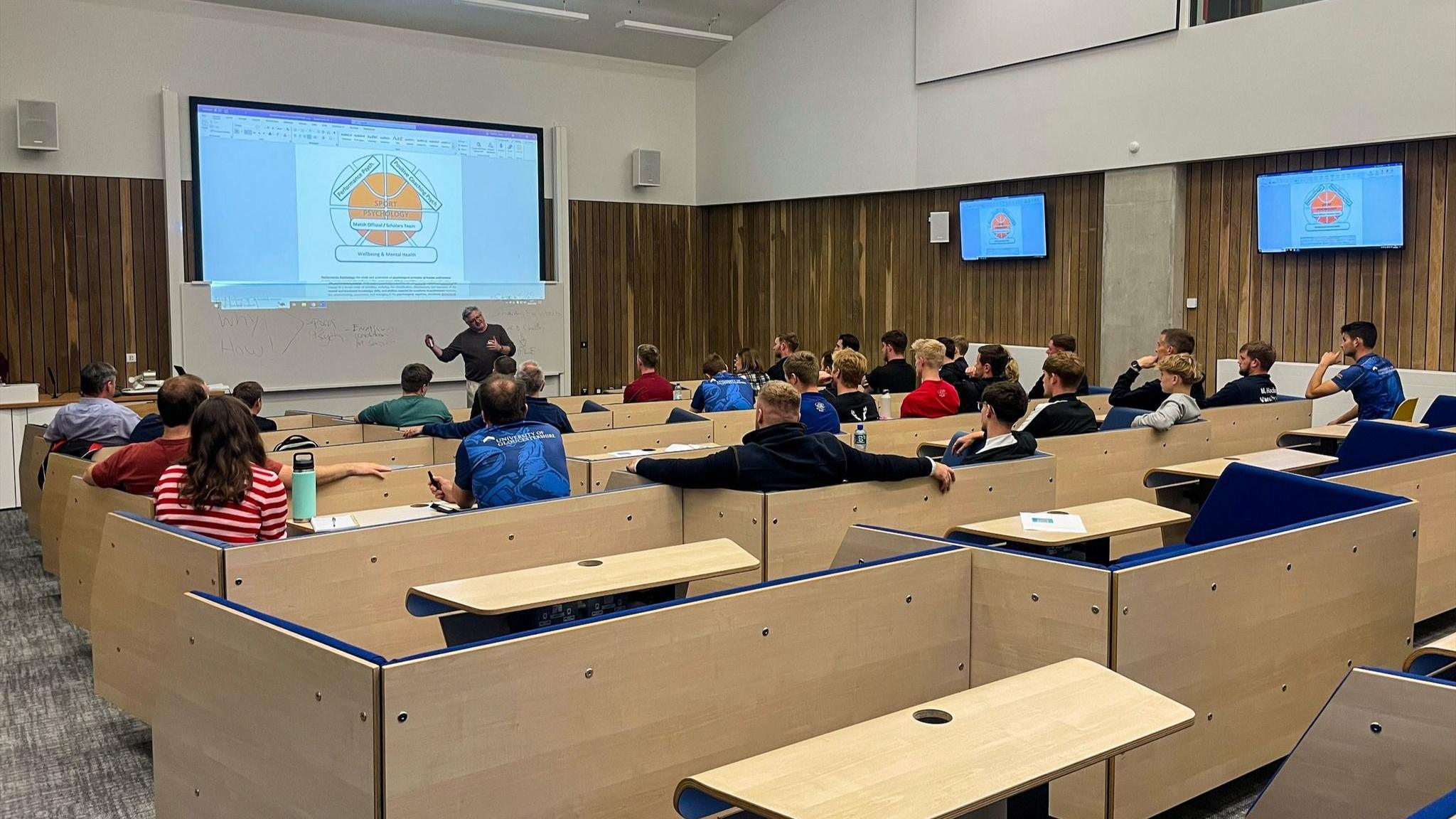 A view of students in a lecture hall from behind with a lecturer standing at the front