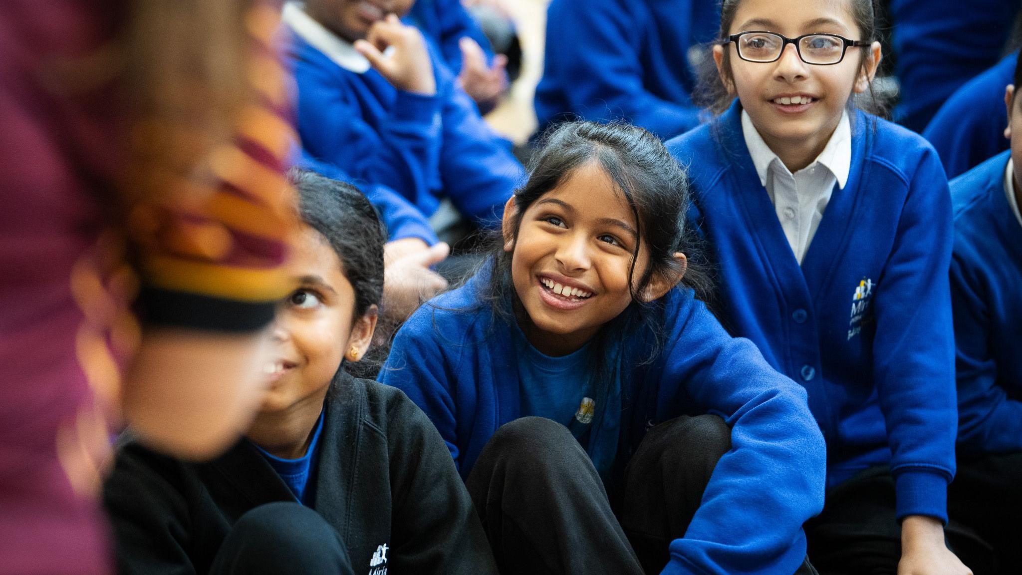 A girl with dark hair is smiling up at a person standing nearby. She is sat next to a girl with dark hair and glassed and another girl who is wearing a black jumper over a blue school sweatshirt.