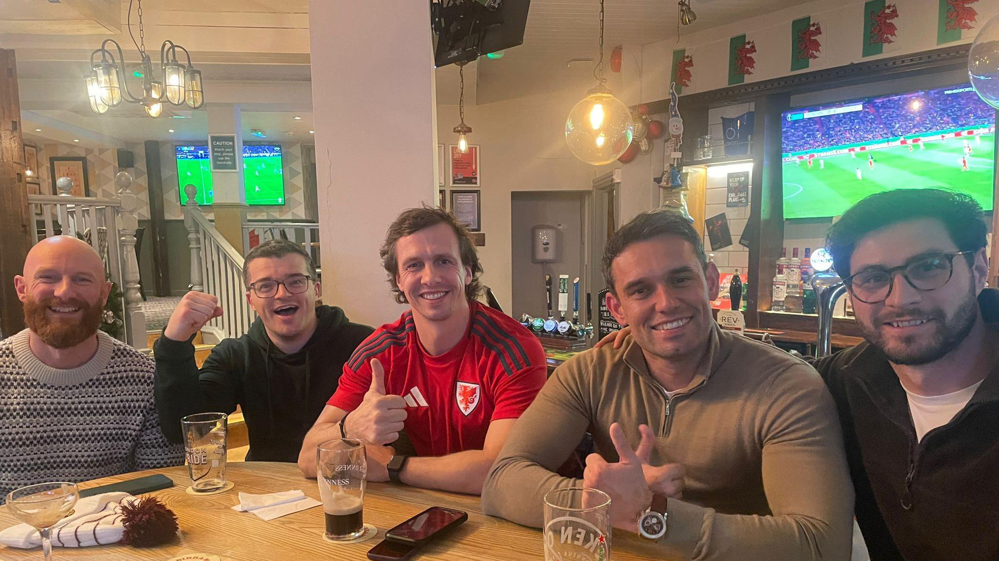 Picture of five men sat around a pub table in Cardiff with near empty pint glasses and mobile phones on the table in front of them. All smiling, one wears a Wales top and holds his thumb up while welsh flags can be seen in the background with TV screens showing the game. 