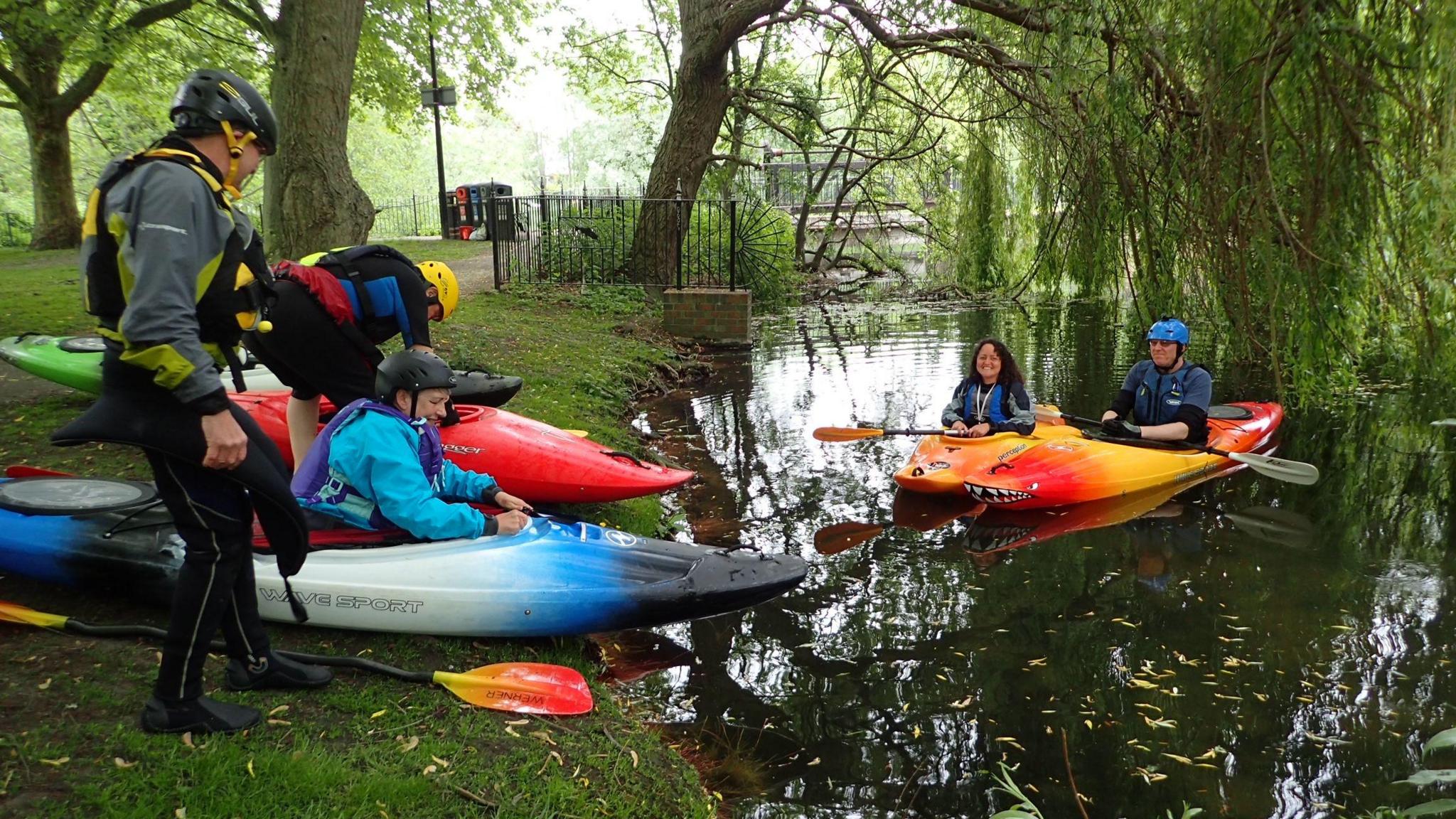 Five people in or getting into canoes on a river. Two of them are already on the water and three are on the grass verge next to it. There is a large tree behind them with sweeping branches falling down towards the river.