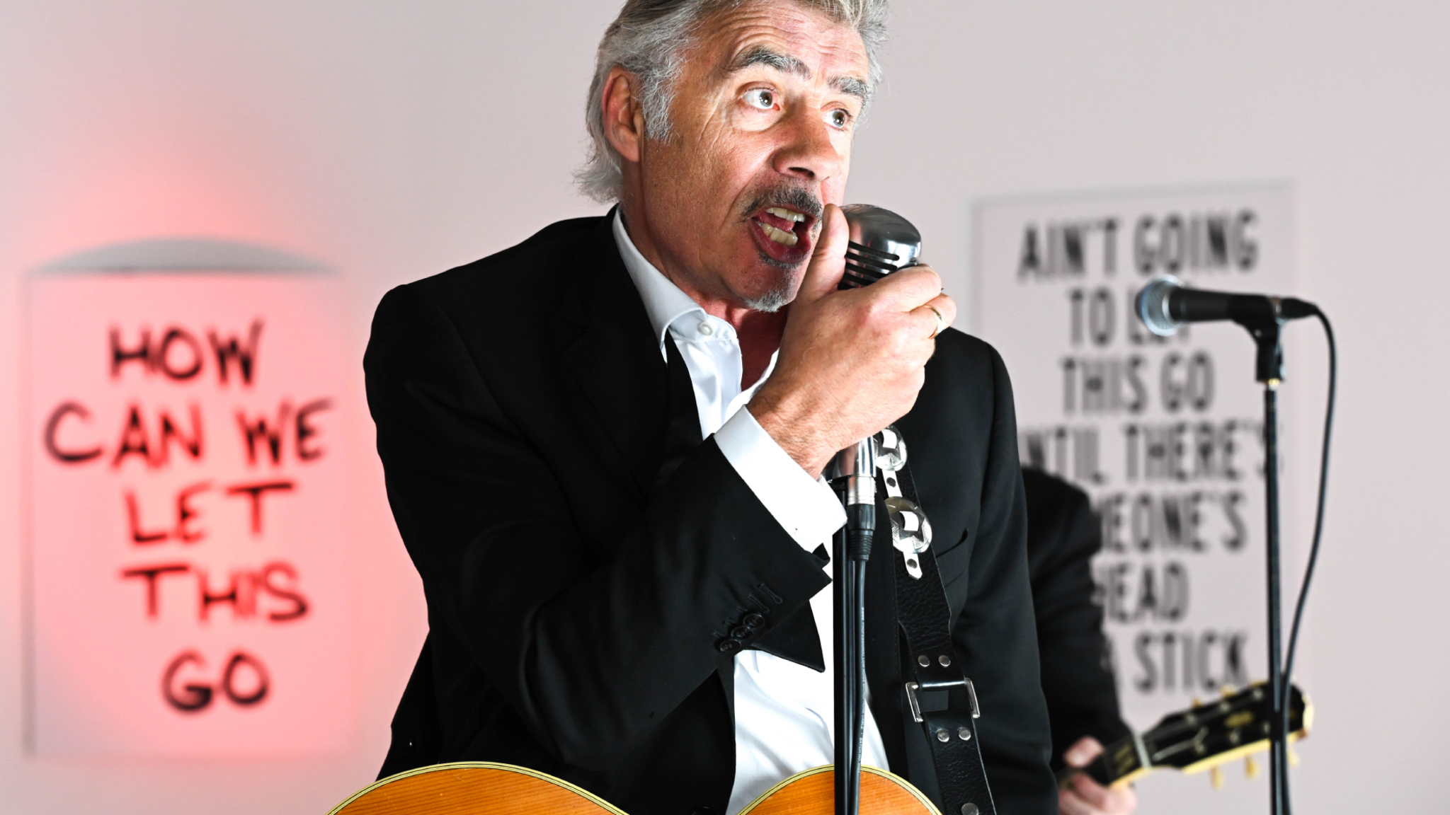 Glen Matlock sings into a microphone, with a guitar around his neck, with slogans on the wall behind him, including 'How can we let this go'