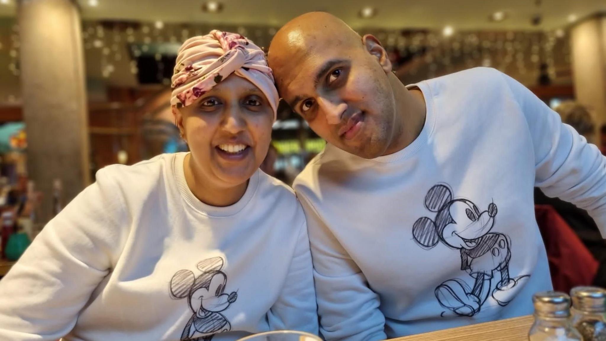 Priyanka Pandya-Bhatt with her husband, Manoj Priyanka, sitting at a table, wearing white tops with Mickey Mouse on Priynaka, has a headscarf on and Manoy has a bald head. They are in a restaurant, with salt and pepper bottles on the table. 