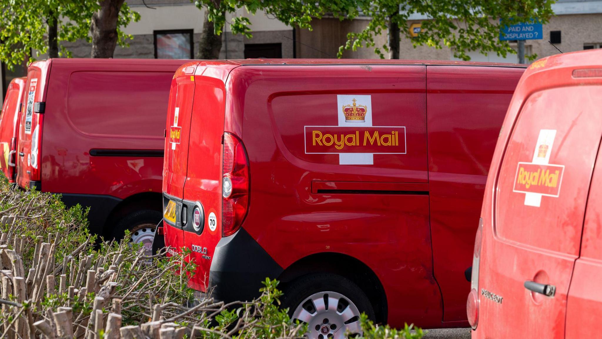 Three Royal Mail vans in a row in a car park. They are red and have the logo on. We can see some bushes around them and a tree behind them. 