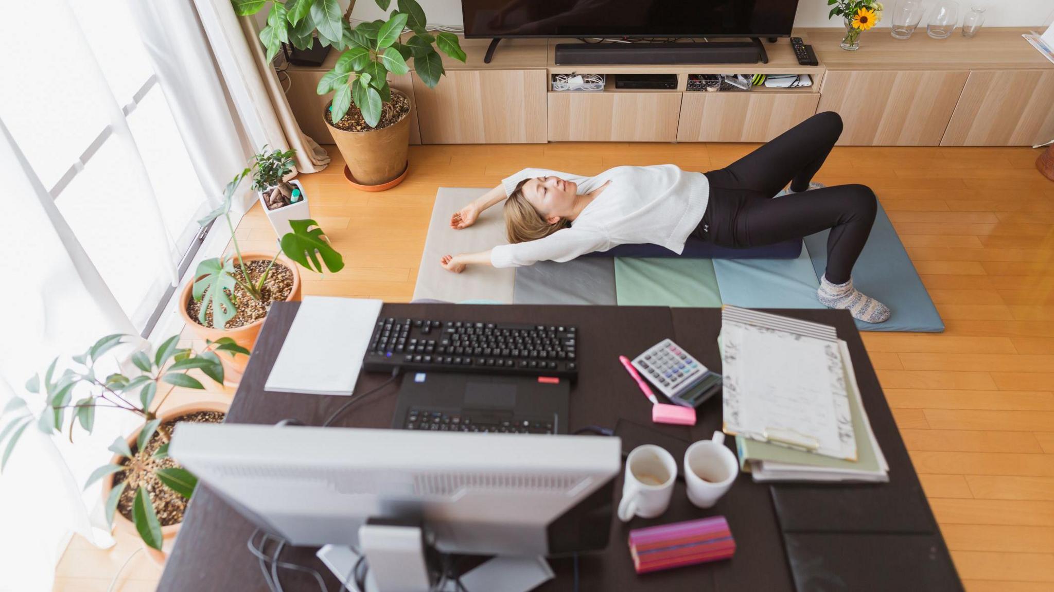 A woman working from home and exercising on a yoga mat in a living space.  Next to her is a home office setup with a desk, computer, papers, and mugs. The room has wooden floors and plants.