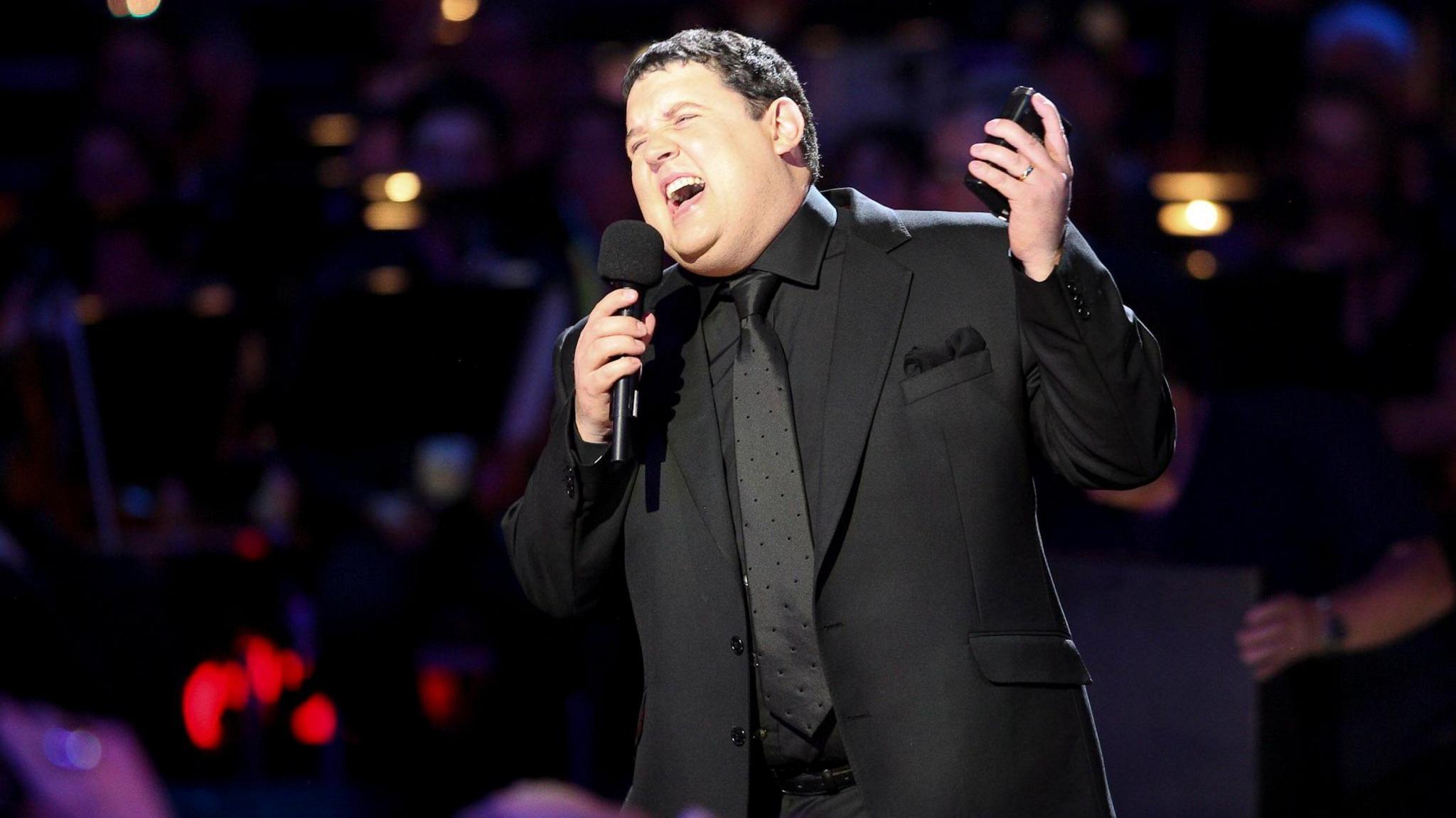 Peter Kay wearing a black suit preforming a gig, holding a microphone and laughing 