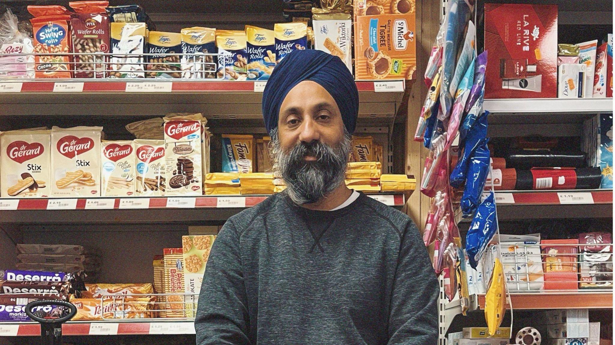 A Sikh corner shop owner in a grey sweatshirt and blue turban, with a grey beard standing in front of shelves containing cakes and biscuits