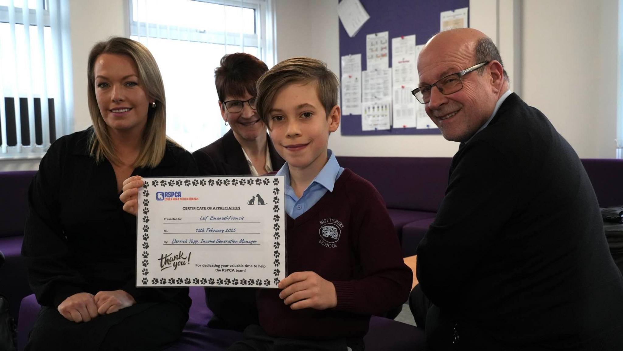 A young boy holds up a certificate from the RSPCA with three adults surrounding him. All of them are smiling at the camera.