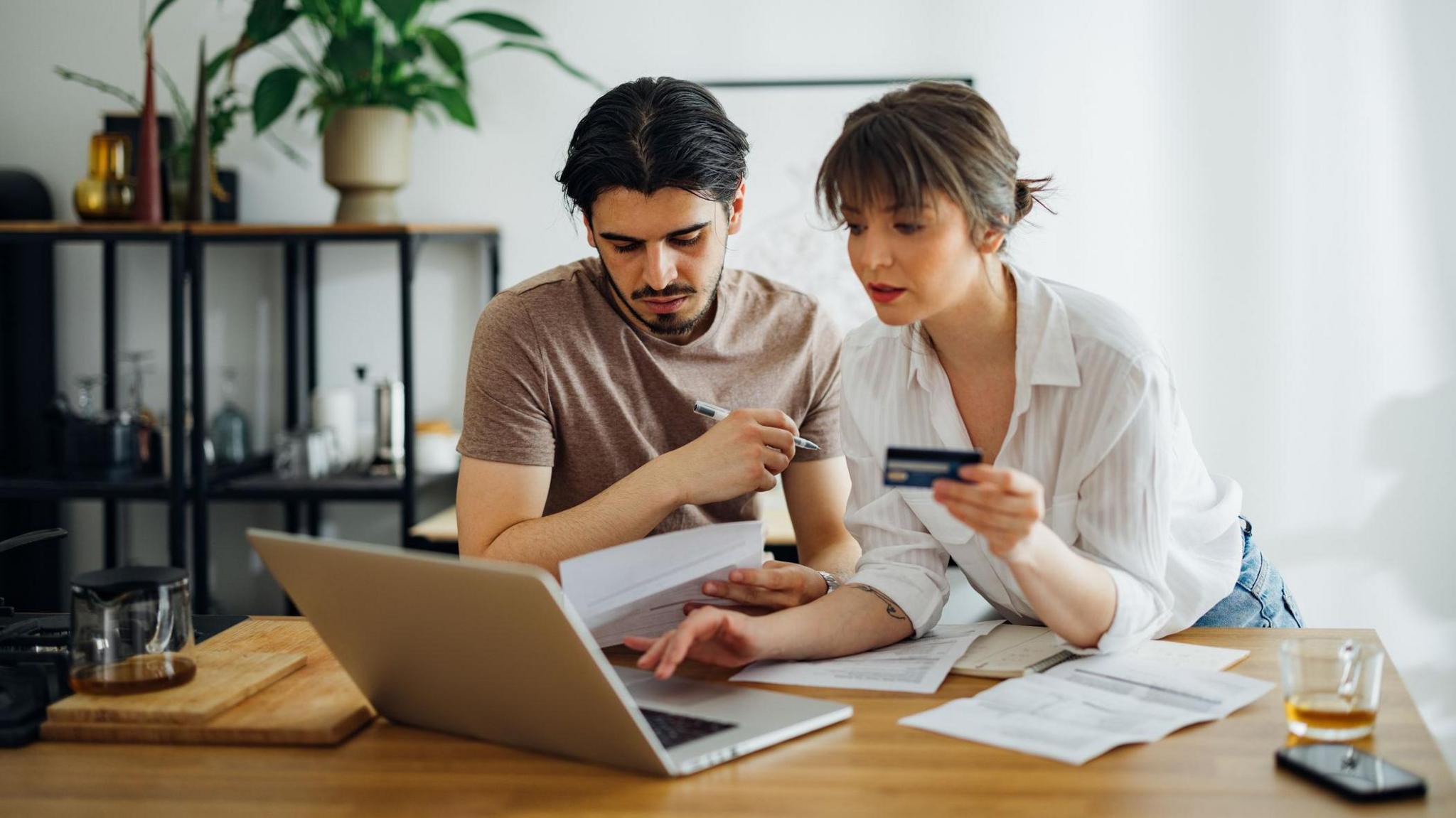 People look at a laptop holding papers and bank payment card - stock shot