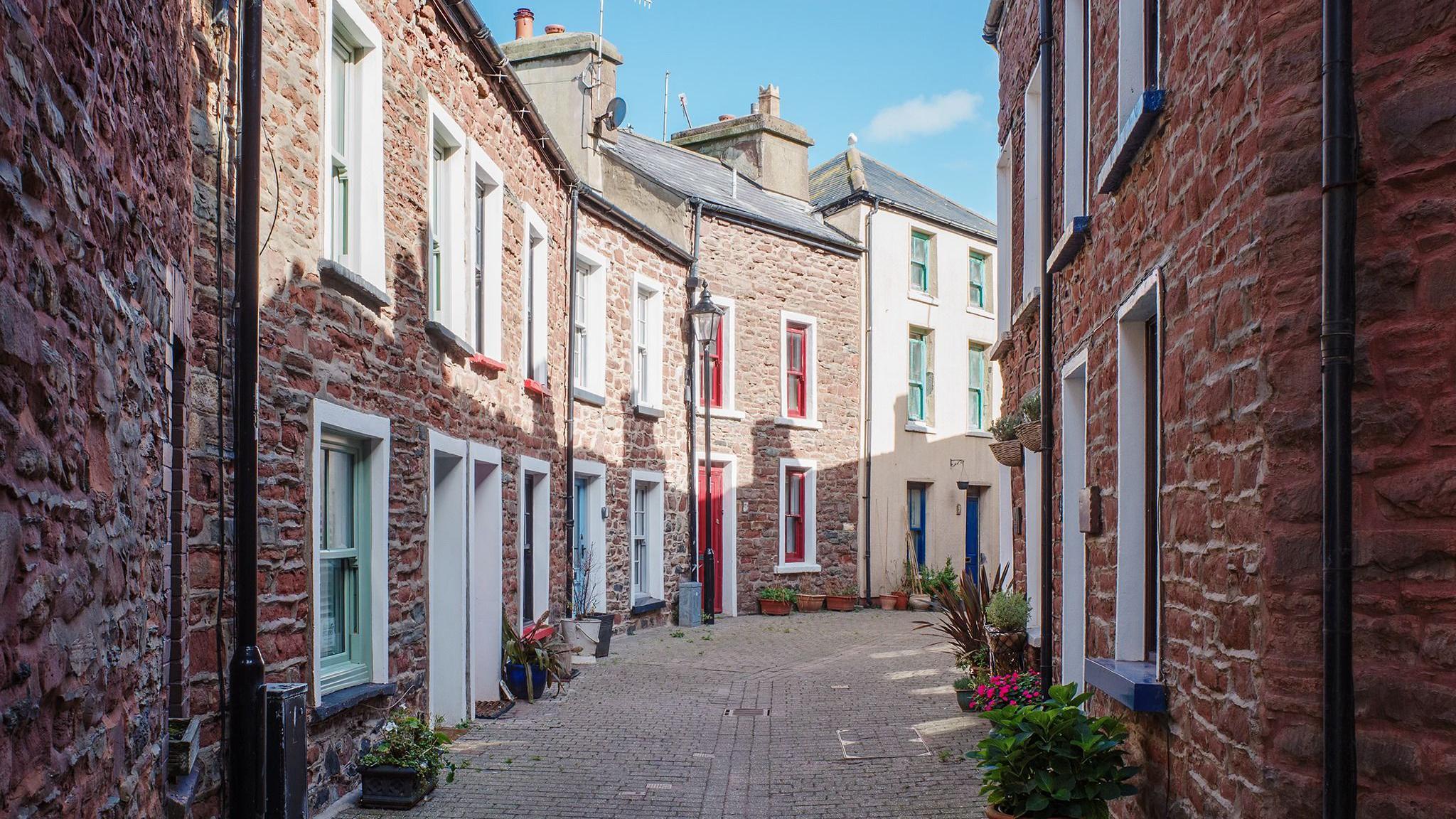 The view of an alleyway between two rows row of quaint red brick cottages. They have brightly coloured window frames and the sky is blue.