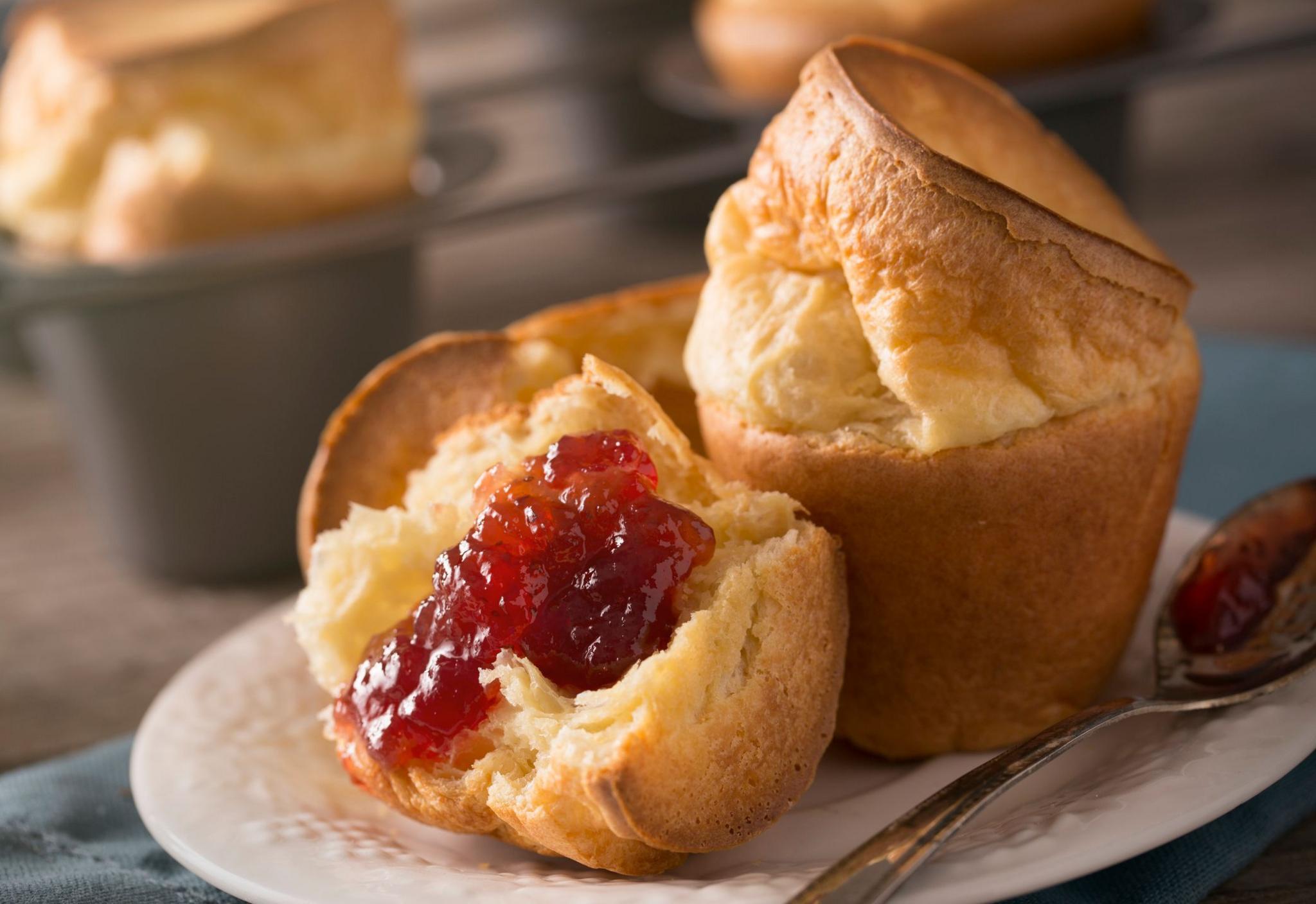 a plate with a couple of yorkshire puddings, one has been spread with strawberry jam as a spoon rests on the side covered in jam