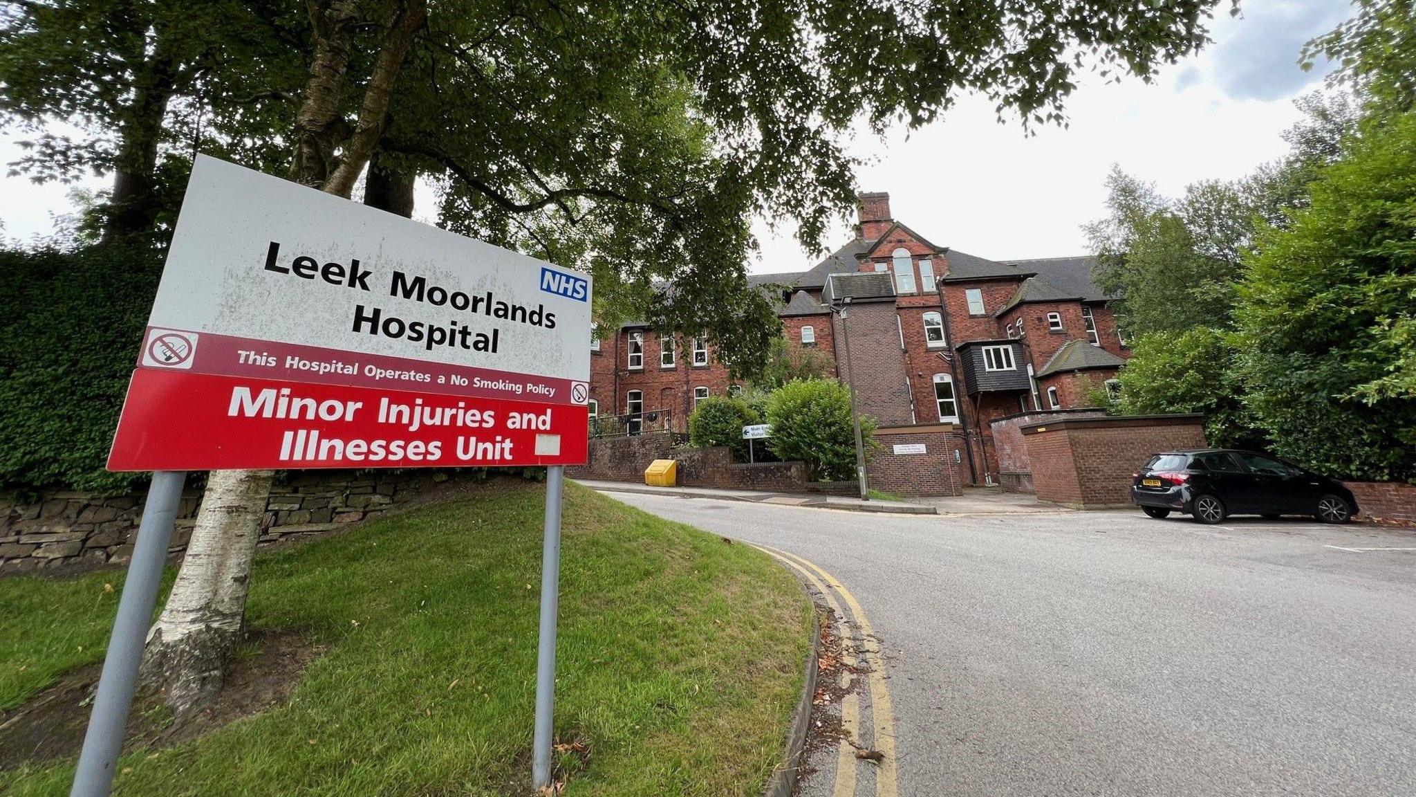 A red and white sign under a tree on the left says "Leek Moorlands Hospital - Minor Injuries and Illnesses Unit". A brick building with walls around it can be seen in the background.
