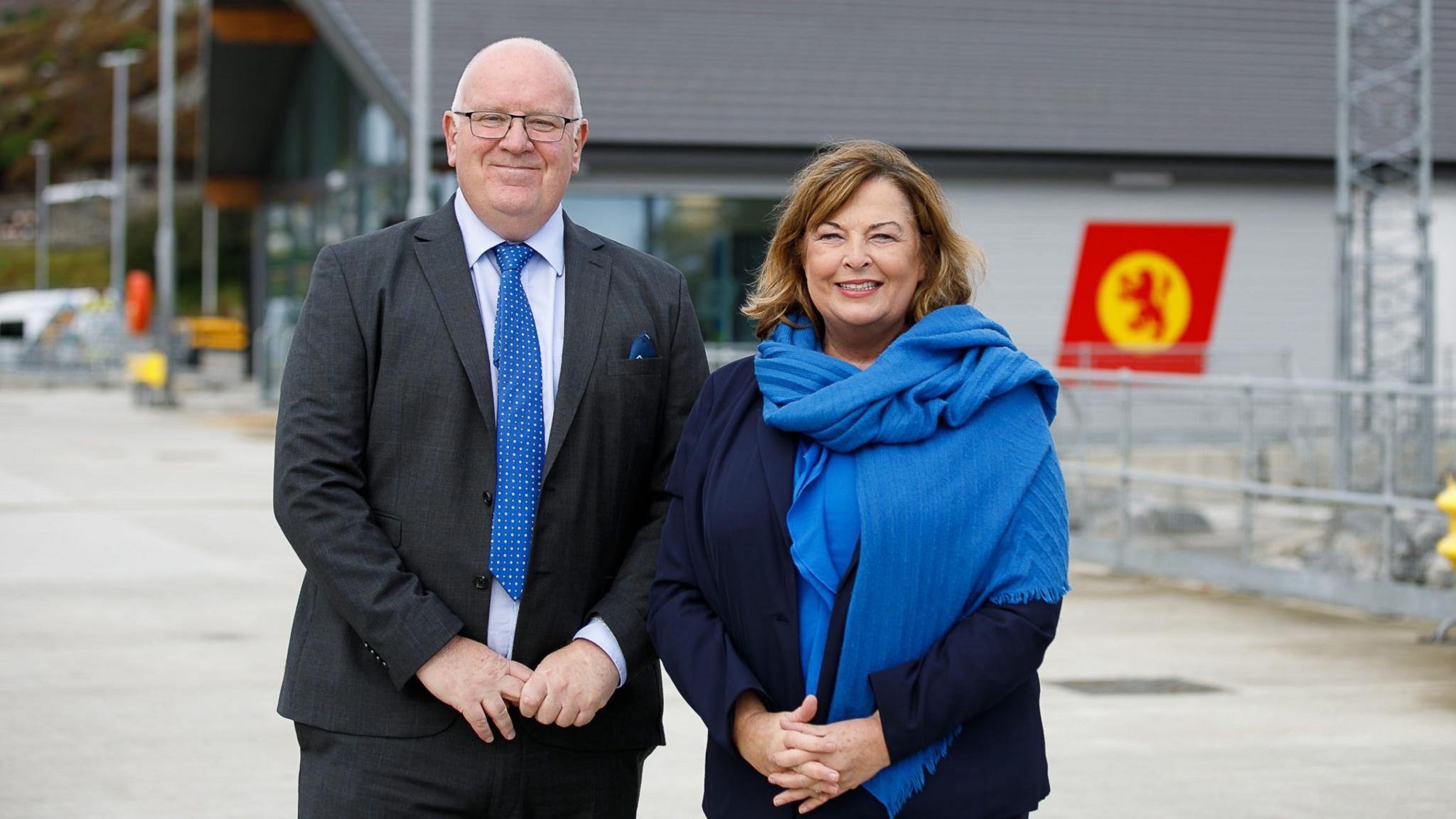 A balding man in a dark suit and blue tie stands next to a woman with a blue shawl at a harbour with the CalMac logo in the background