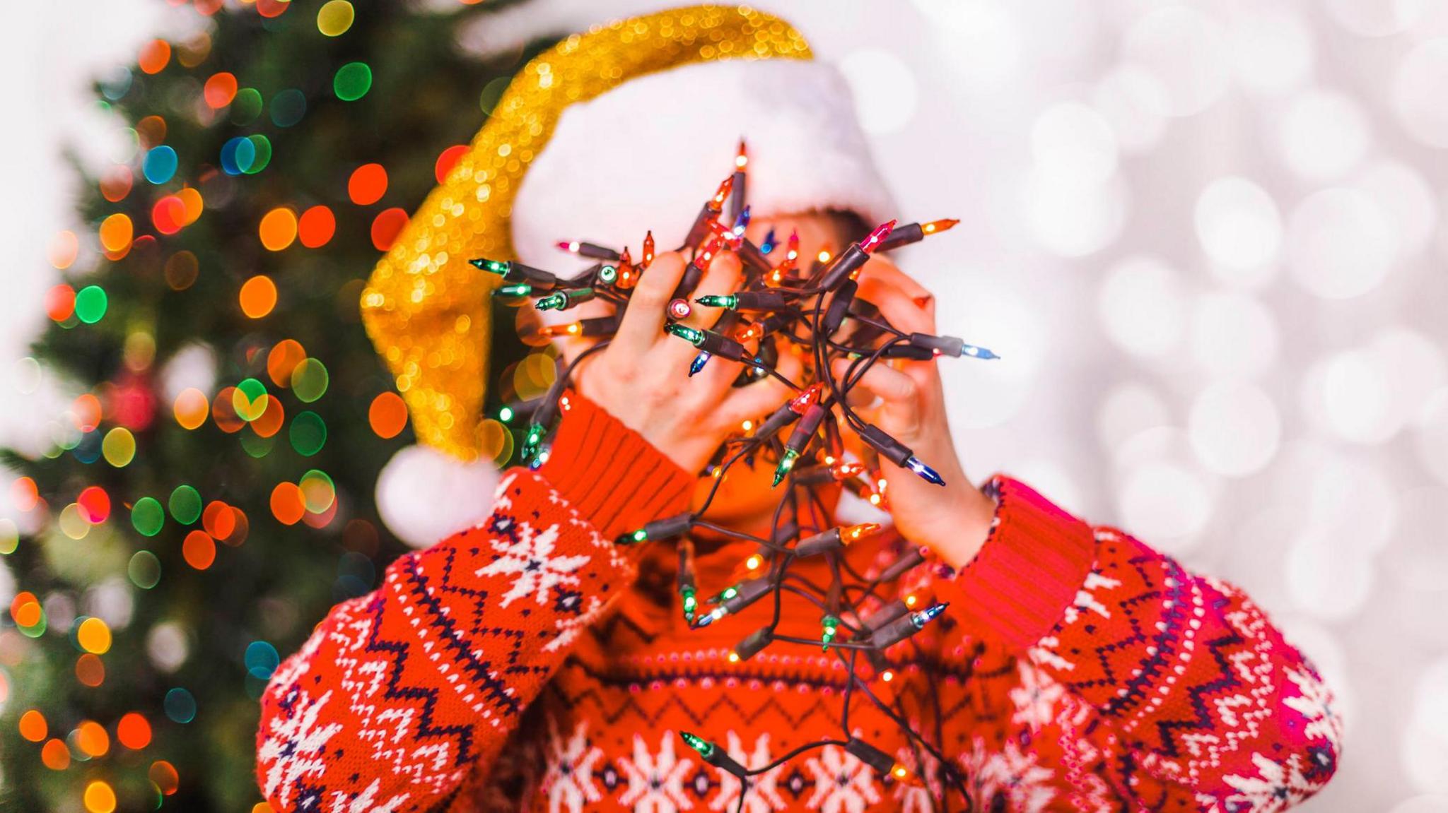 Child wearing a festive red sweater with white snowflake patterns and a gold-trimmed Santa hat, holding a tangled string of colourful Christmas lights in front of their face. A blurred Christmas tree with multi-coloured lights is visible in the background.