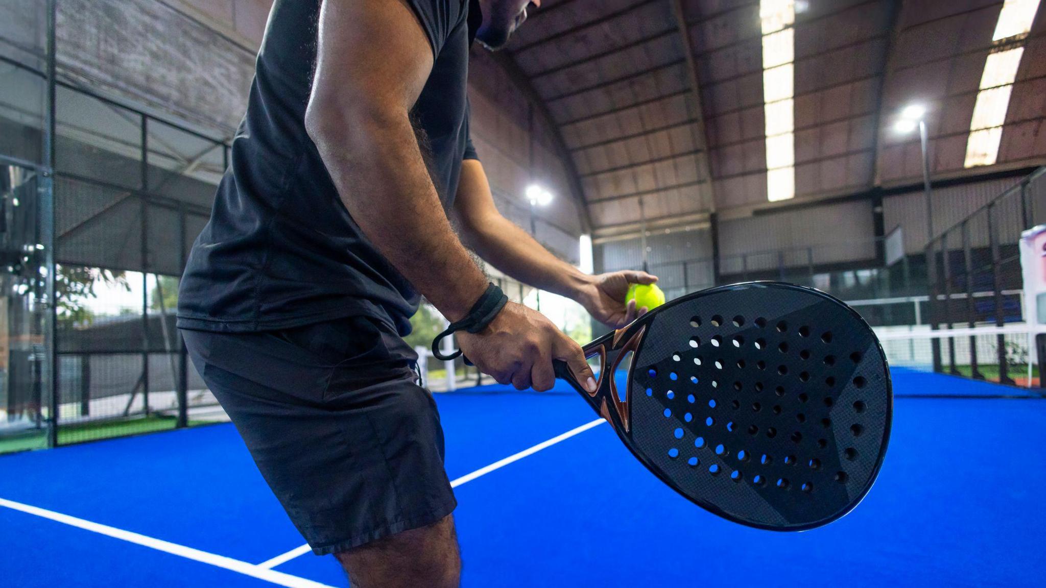 A person holds a padel racquet and ball, with a blue court seen in the background