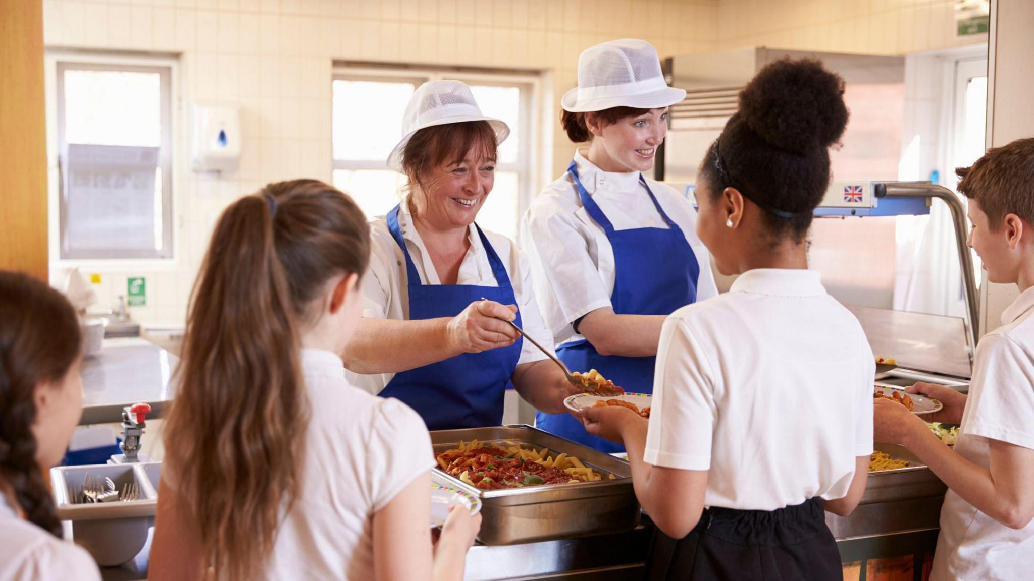 Two women serving children food in a school cafeteria.