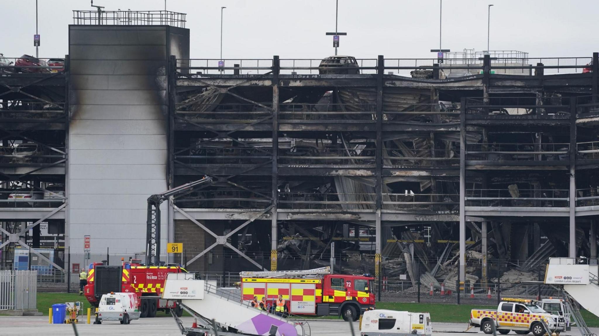 The burnt out and partially collapsed car park building. Fire engines are parked in front of it. It is a grey building, but now mostly scorched black.