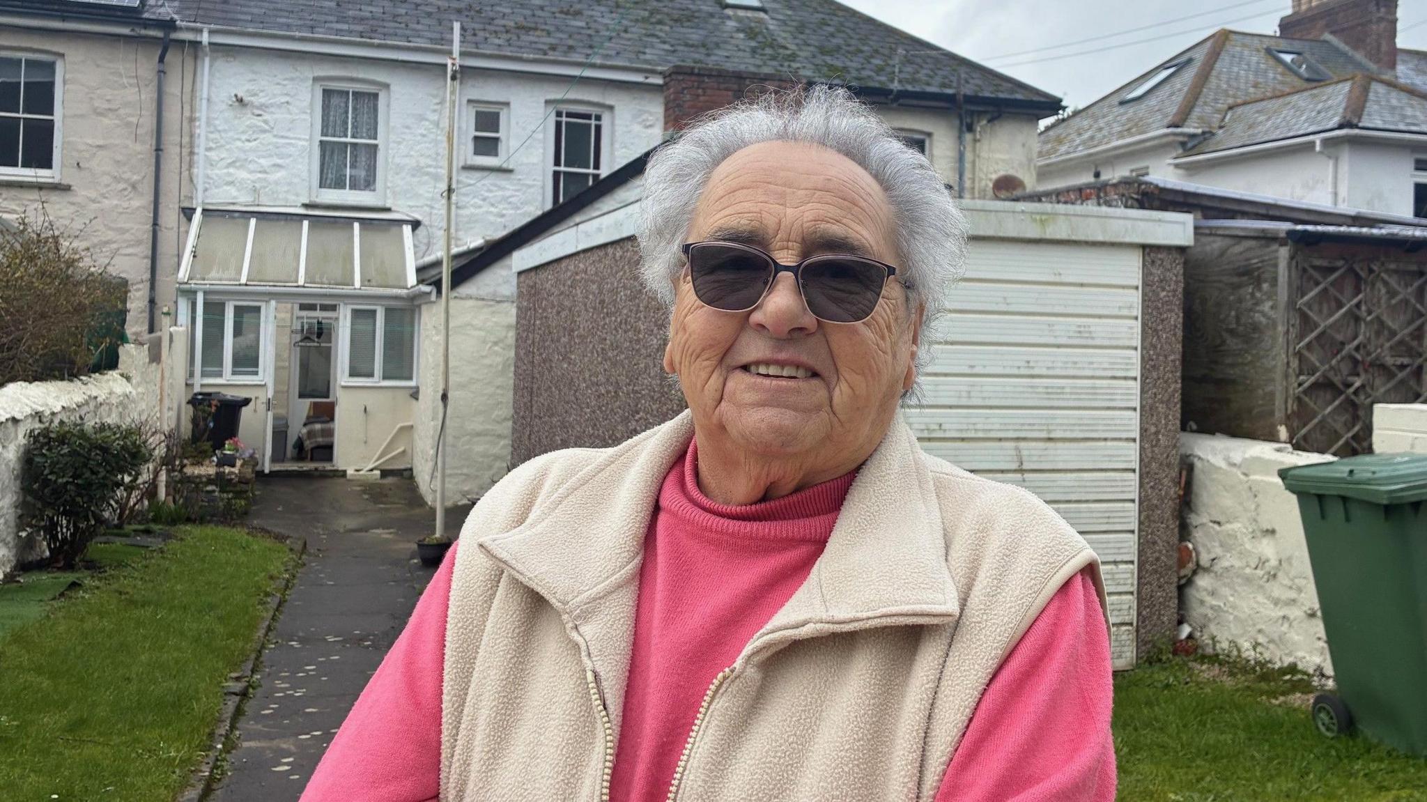 A woman wearing dark glasses smiles at the camera , she is standing in her back garden which is right next to the building site.