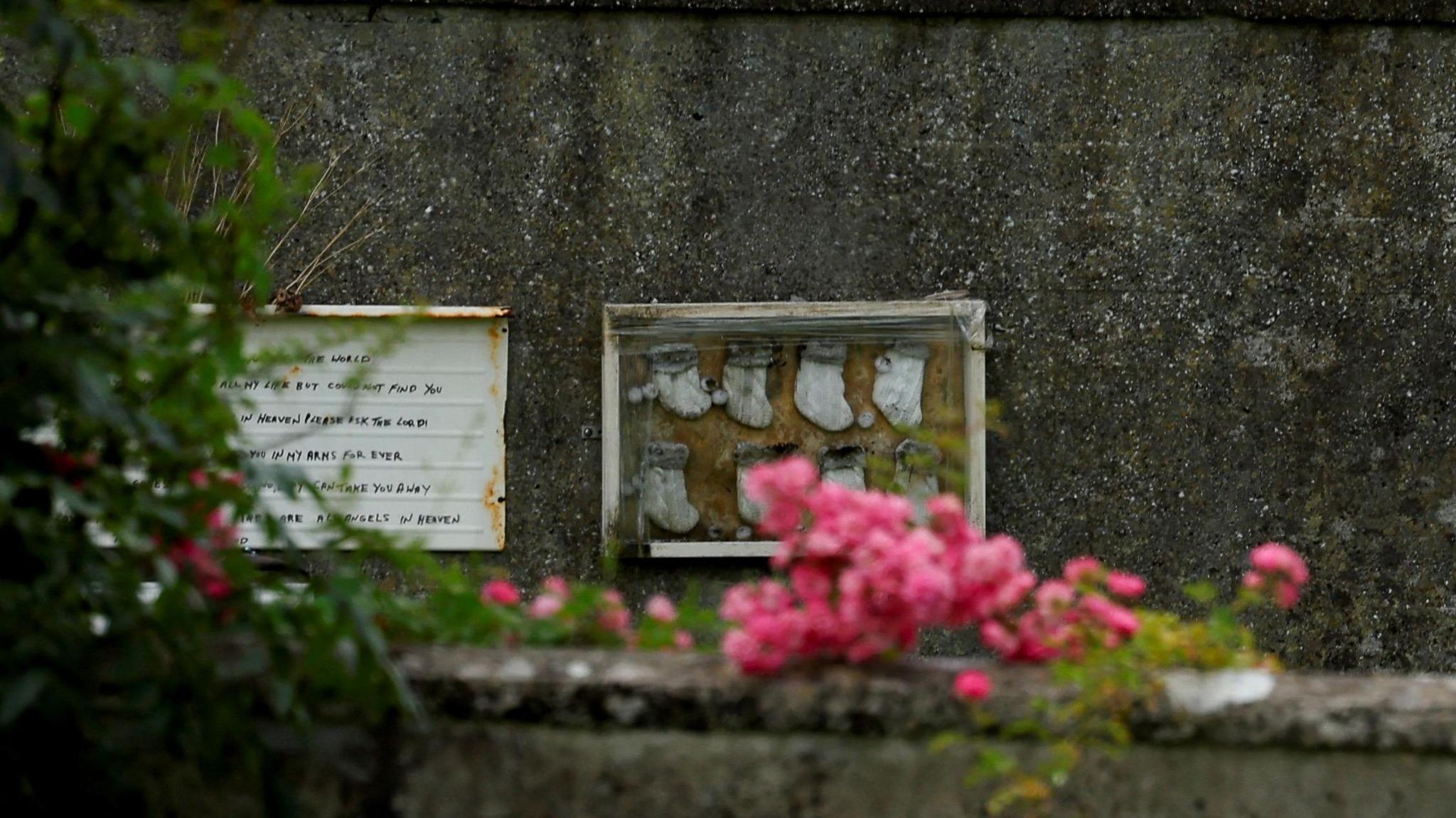 A memorial with a number of small children's socks is attached to a stone wall. In the foreground is pink flowers.