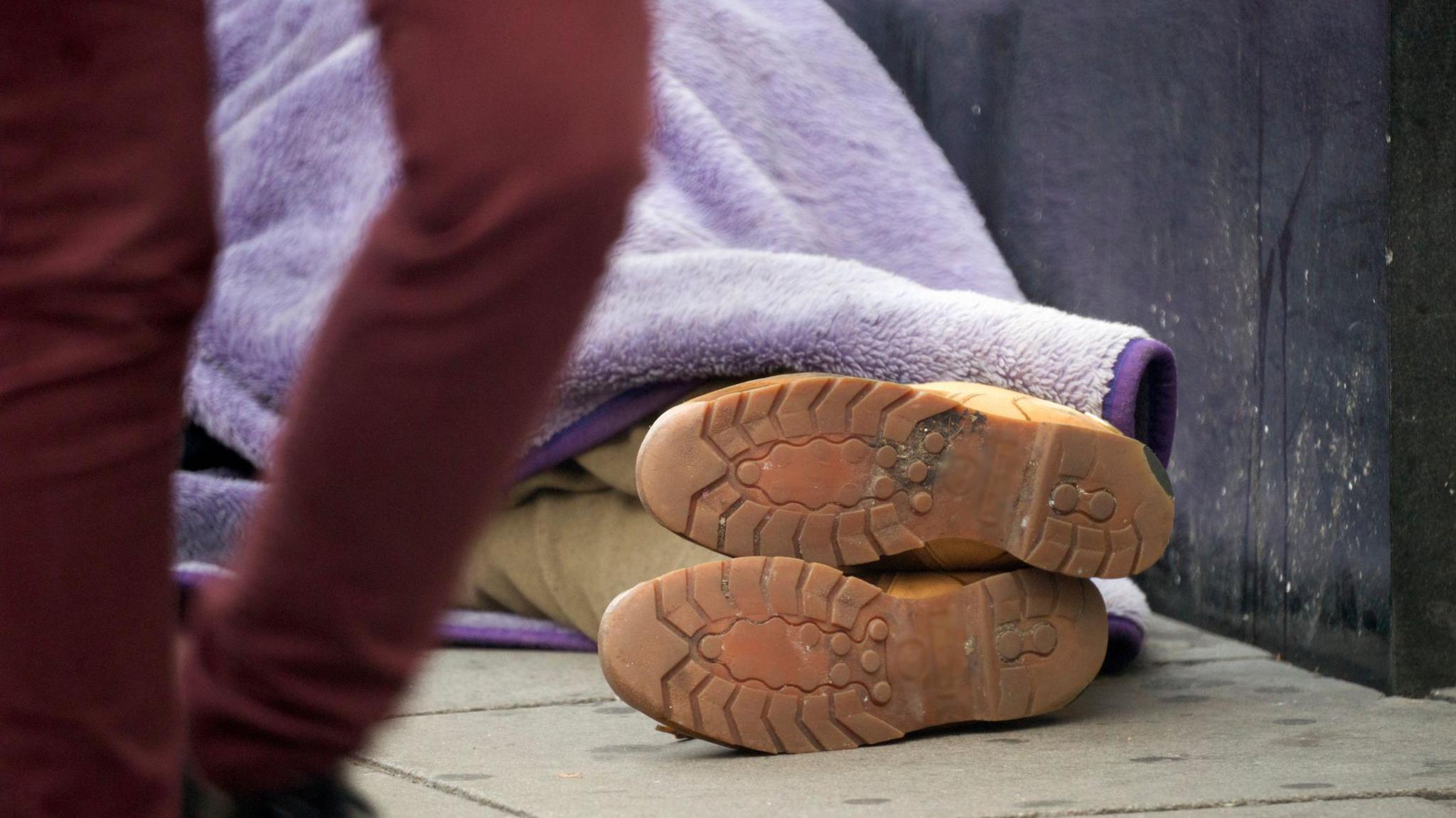 A homeless person is sleeping on the pavement, they are lying on the floor and only the soles of their shoes are in vision, they are a pair of beige rubber soled boots and the person wears beige trousers, with a purple fleece blanket over the top of them, they are lying up against a black wall