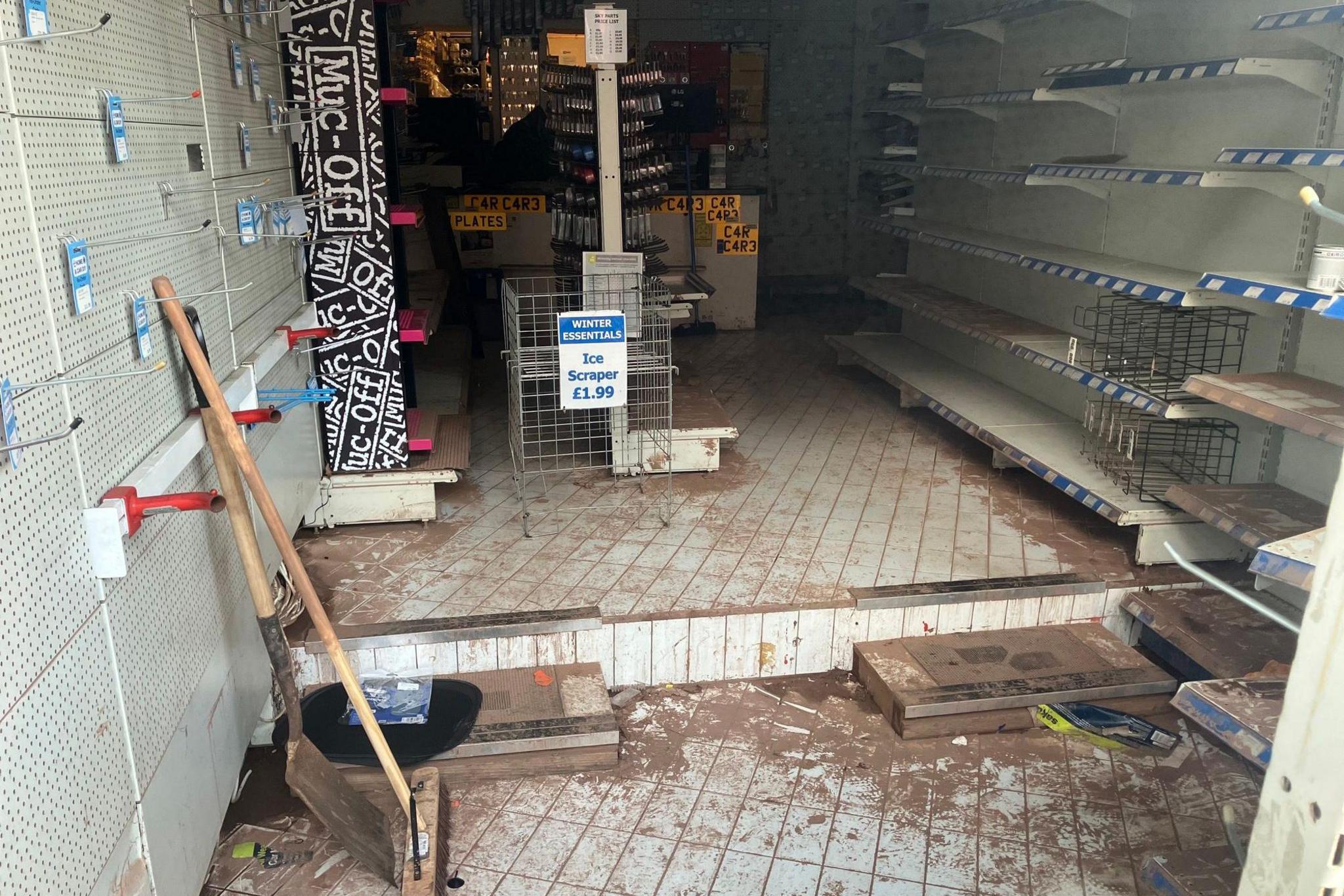 A shop looks empty with all shelves cleared. There is dried mud on the floor and a shovel and a brush leaning on a wall.