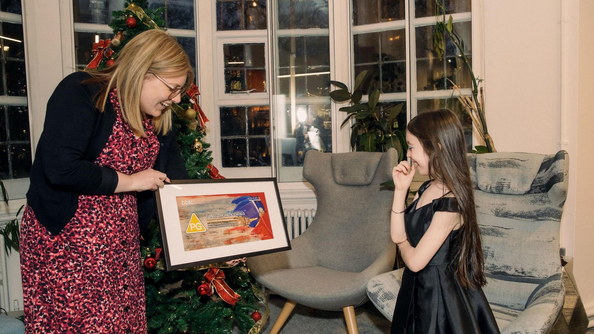 A woman is holding a copy of Ruby's painting signed by the cast of Sonic 3. Ruby is laughing