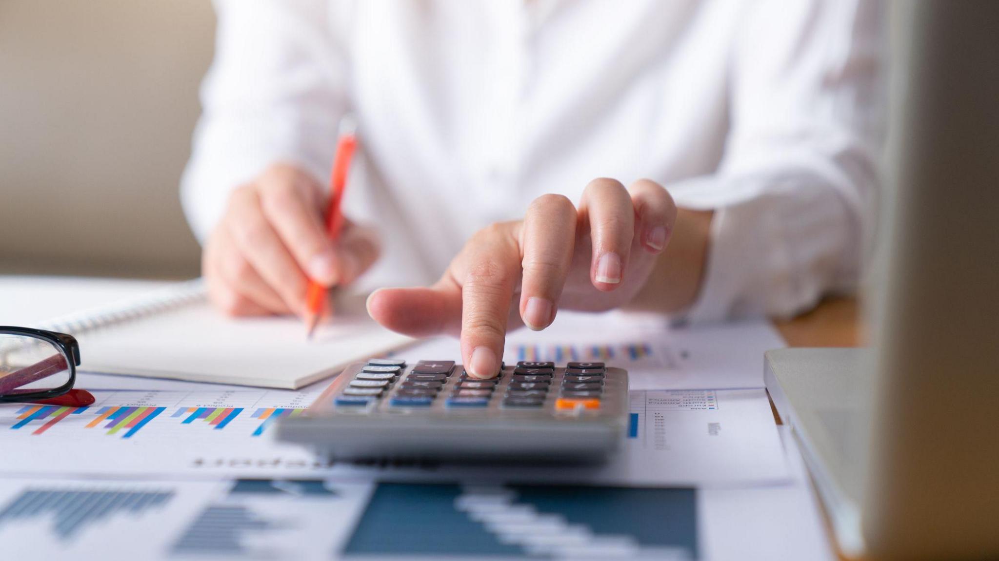 Woman with spreadsheets and calculator in front of her. There is a laptop and glasses on the table. She is wearing a white shirt and holding an orange pencil while using the calculator.
