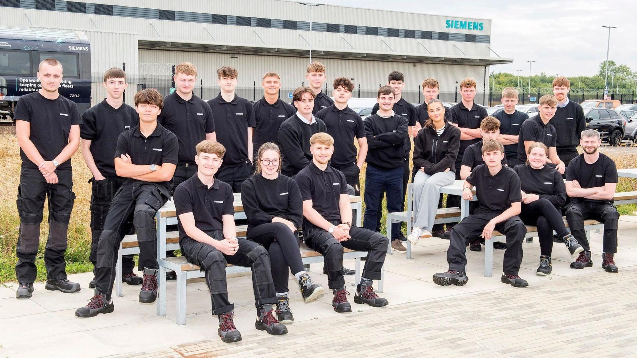 A group comprising 21 young males and three young female in front of a building displaying a Siemens Mobility sign. Some are standing, with other sitting.