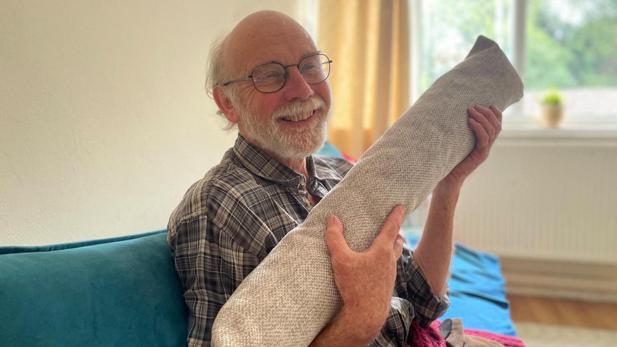 A smiling man hugs a long draught excluder as he sits on a blue sofa