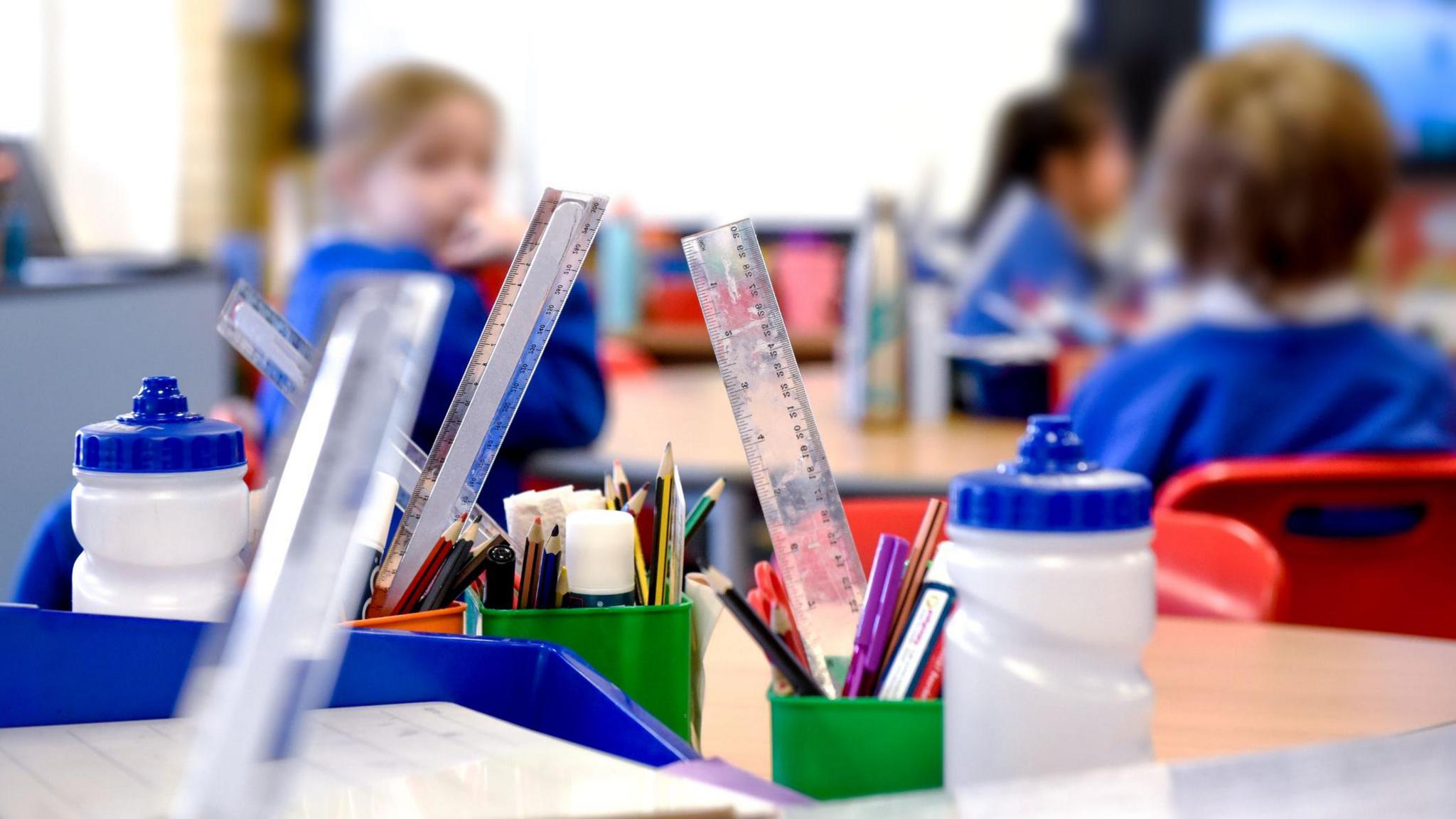 blurred image of children in a primary school classroom with stationary and water bottles in the foreground