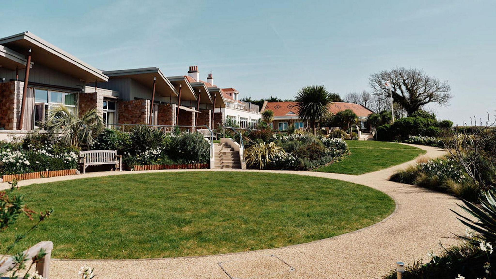 The hospice has units which are single storey and have balconies. There is circle of lawn with a wooden seat overlooking it and a path around it. There are also flower beds and palm trees.