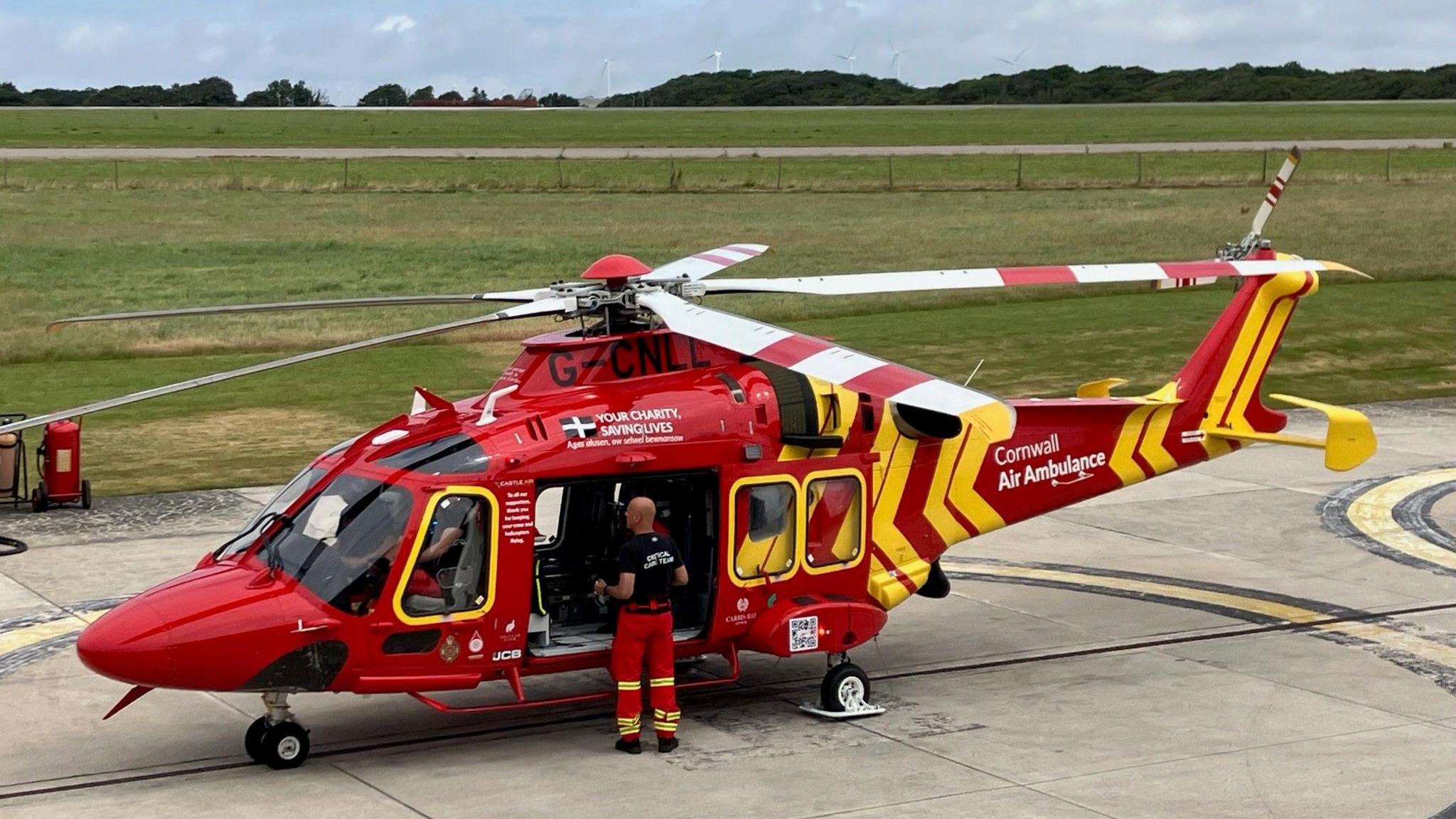 Mick McLachlan is standing in the open doorway of a red air ambulance helicopter, which is parked on the concrete landing pad. An airport runway can be seen in the background across a section of grass.