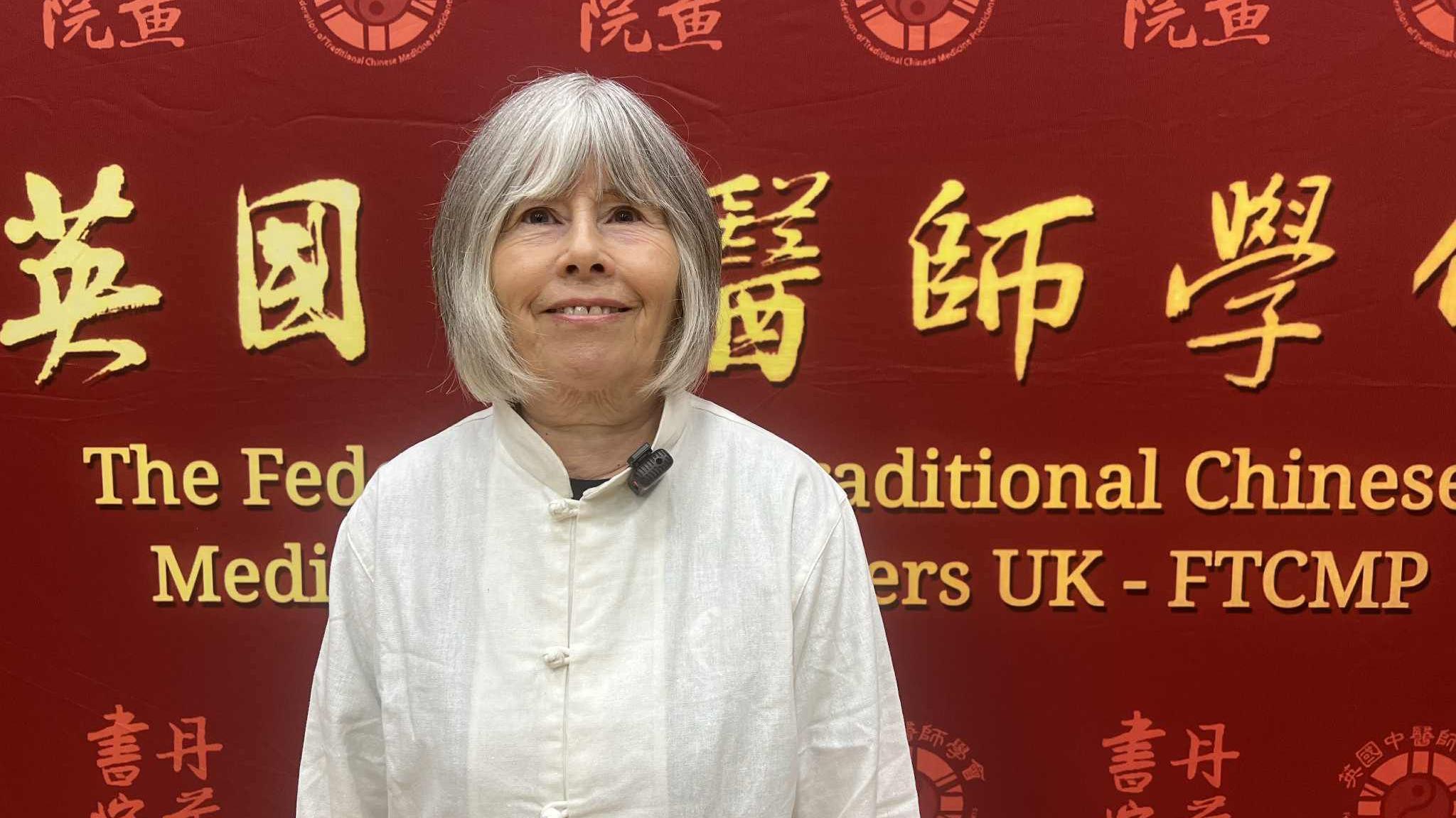 The picture features a woman with white hair wearing white traditional Chinese clothing. Behind her is a wall with large Chinese font, with an English translation below. The wall features Chinese symbols.