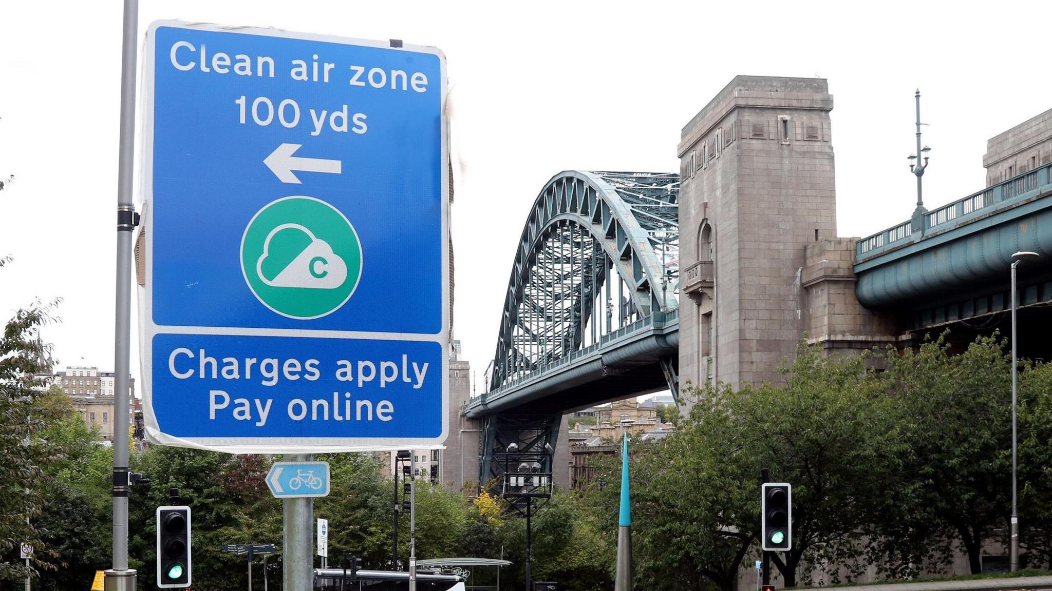 A view of a blue sign reading 'Clean air zone 100 yds' and 'Charges apply pay online'. In the background is a large bridge in Newcastle.