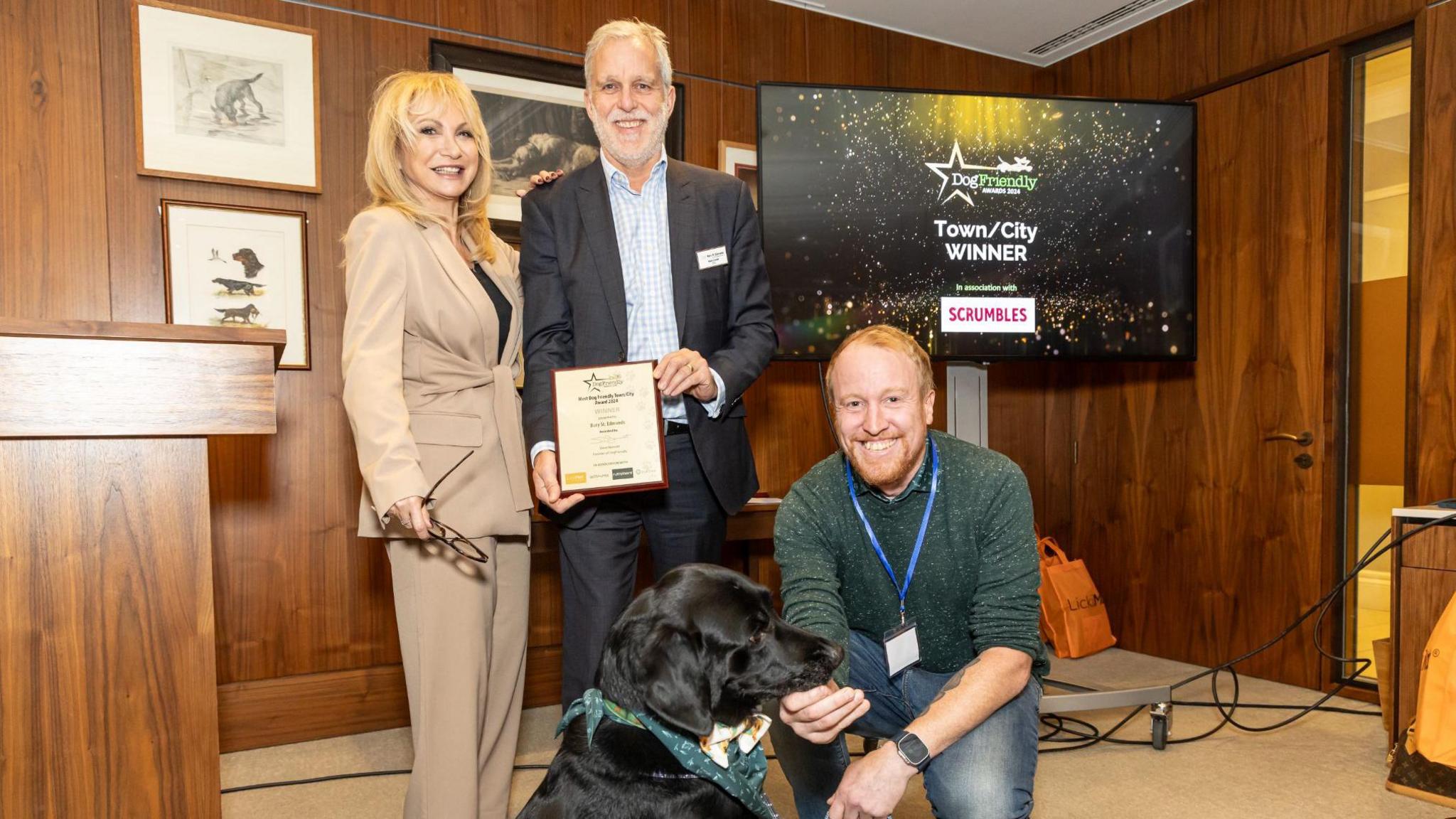 Two men and a woman posing with black dog while holding an award