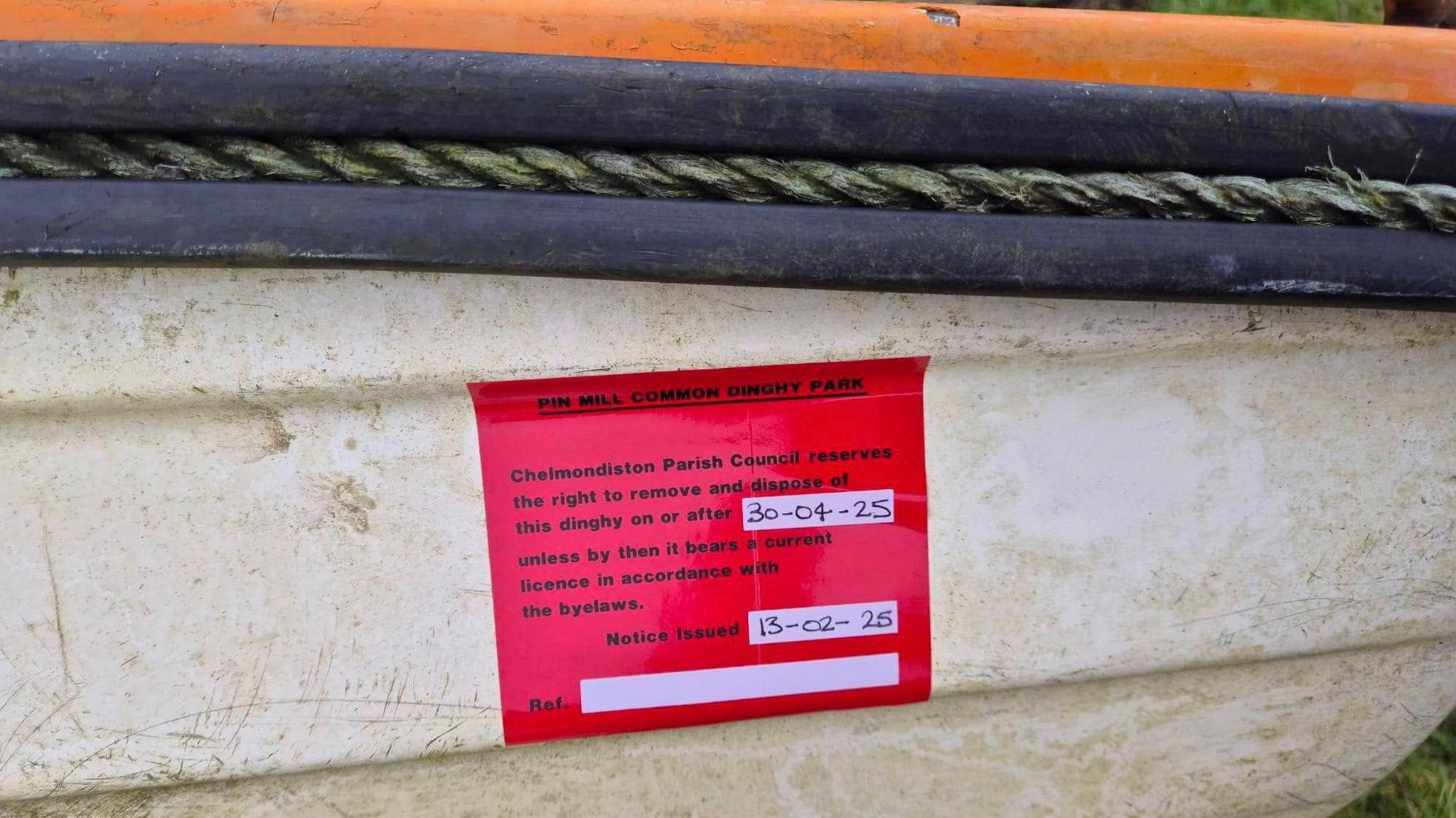 A red plastic notice is stuck to the side of a white plastic dinghy. It is an official council notice that warns the boat owner that the unlicensed boat could be removed after the end of April. A strand of rope runs along the upper part of the dinghy, as part of the rubber fender.    