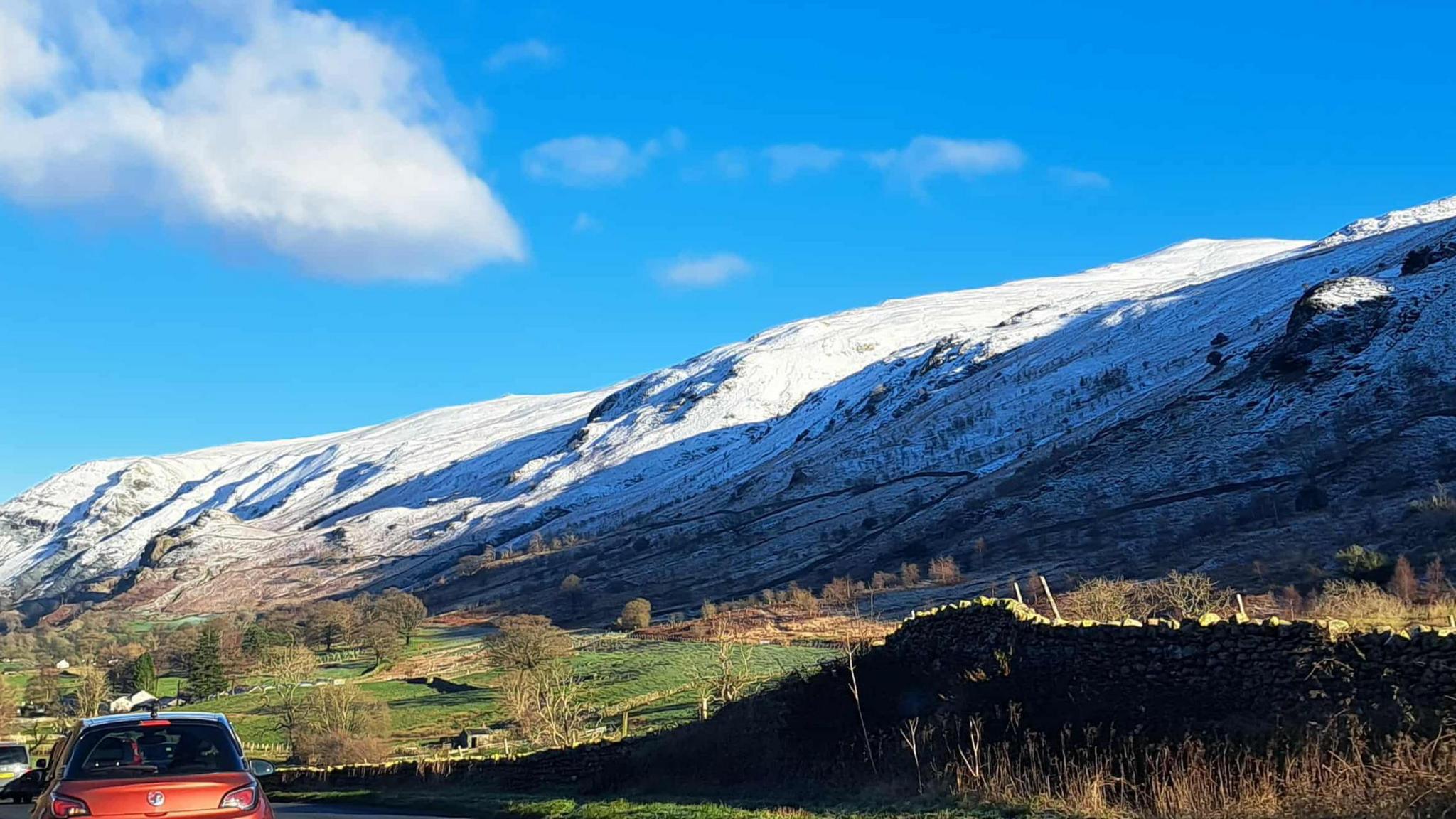 Rounded fells which are covered in snow. The colours transition from a bright blue sky, to white fells then to bright green grass on the ground. There is an orange car driving on the road, near Keswick.