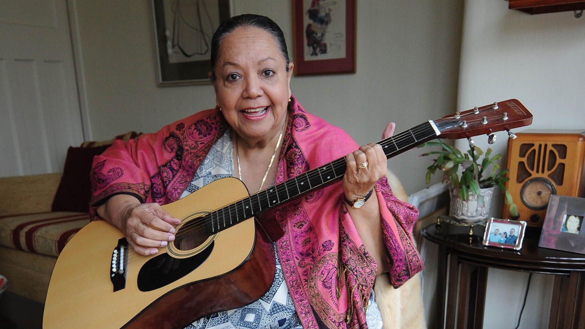 Candy Devine smiling at the camera as she sits on a sofa holding a guitar. She has her hair tied back and is wearing a pink shawl and a black and white patterned dress.