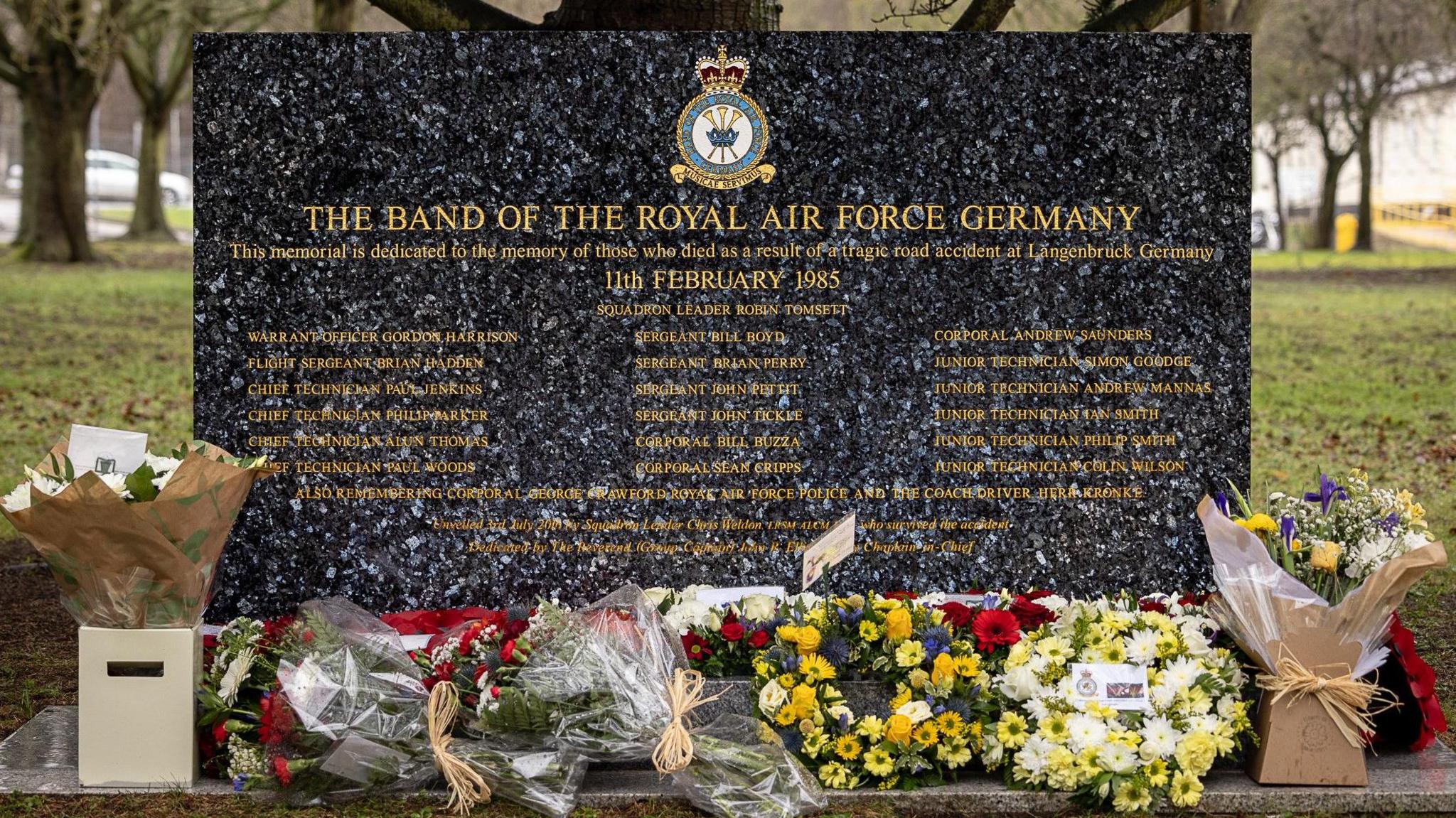 A black marble memorial stone with gold text inscribing the names of the 21 people killed in the crash. Flowers have been placed in front of the stone.