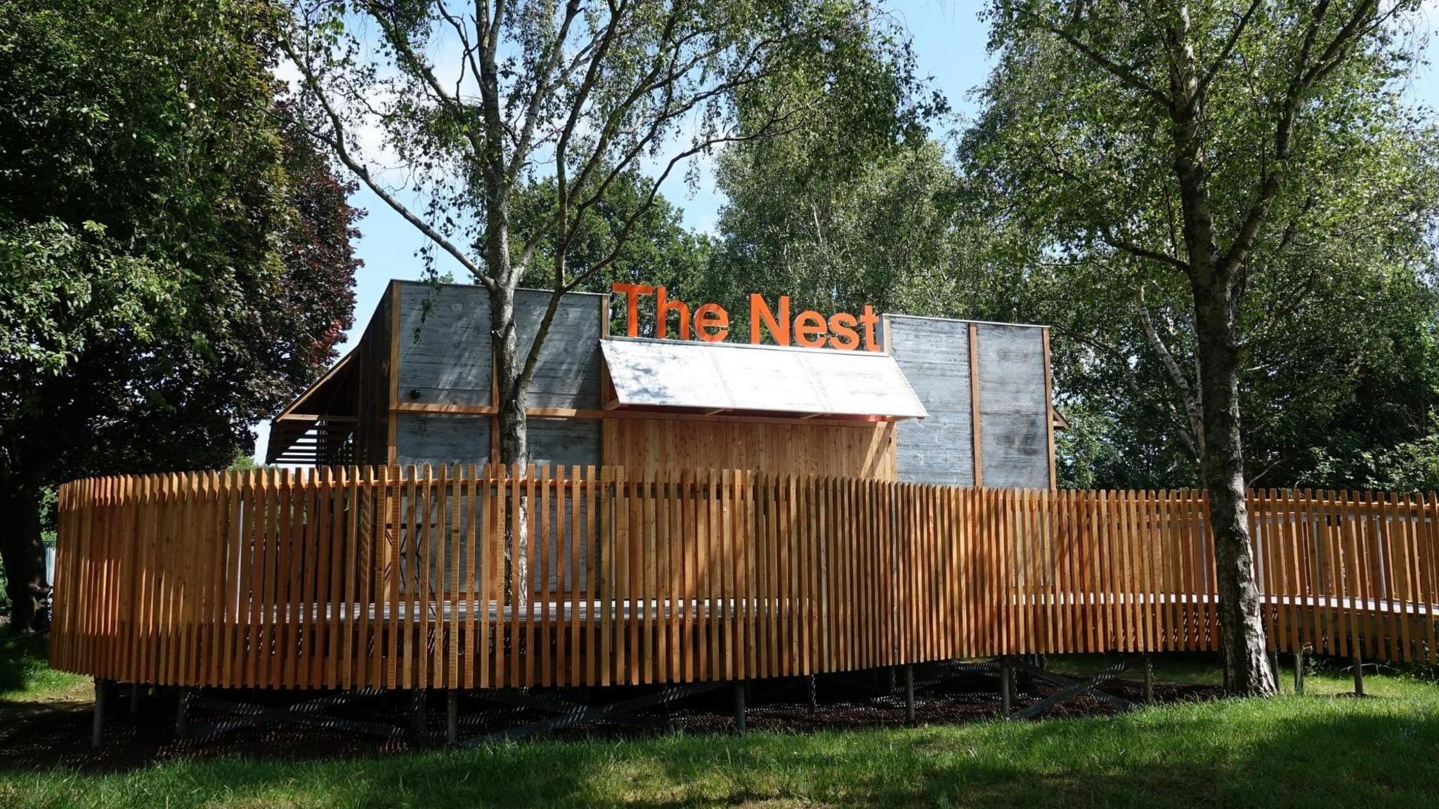 An exterior image of the Nest in Chichester, a performance area set in parkland and surrounded by a tall brown fence.