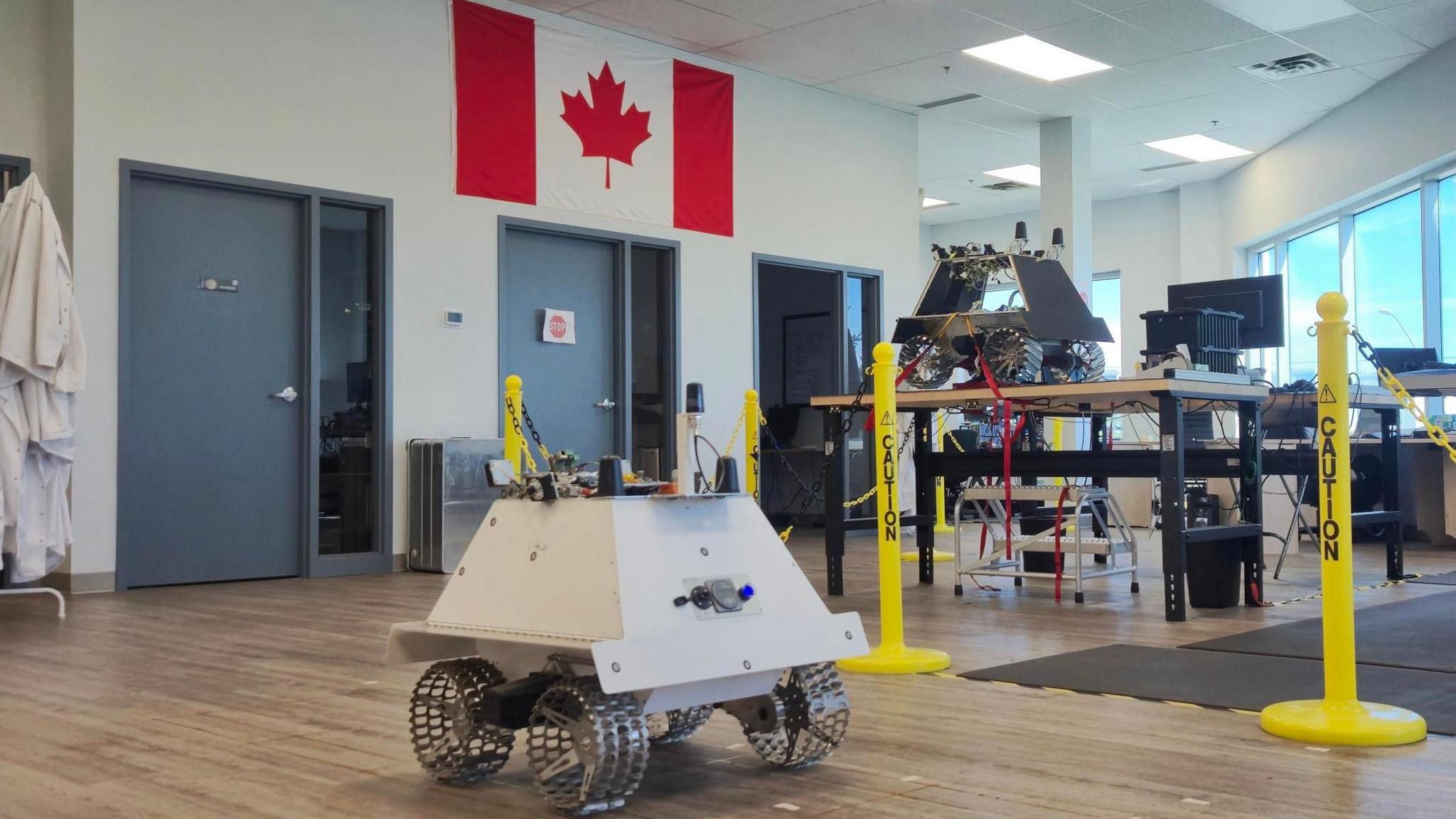 Two prototypes of the Moon rover, one white one in the foreground and the grey one on the table, with the Canadian flag in the background.
