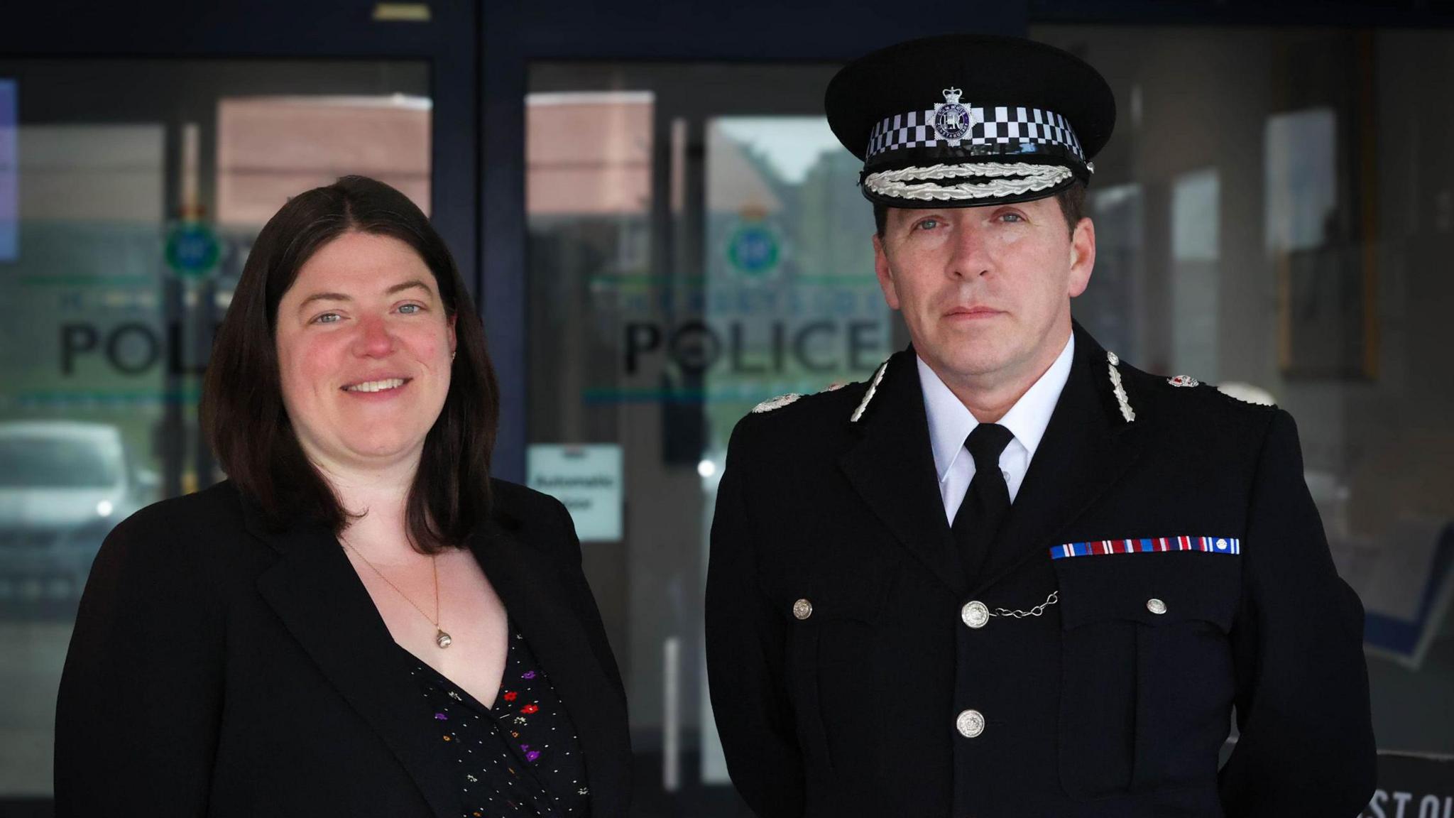 Emily Spurrell has mid-length dark hair and is wearing a black patterned dress, a black suit jacket and a pendant necklace. Rob Carden is wearing a police uniform and hat and he has short, dark hair. Both are standing outside Merseyside Police headquarters and they are both looking directly at the camera.