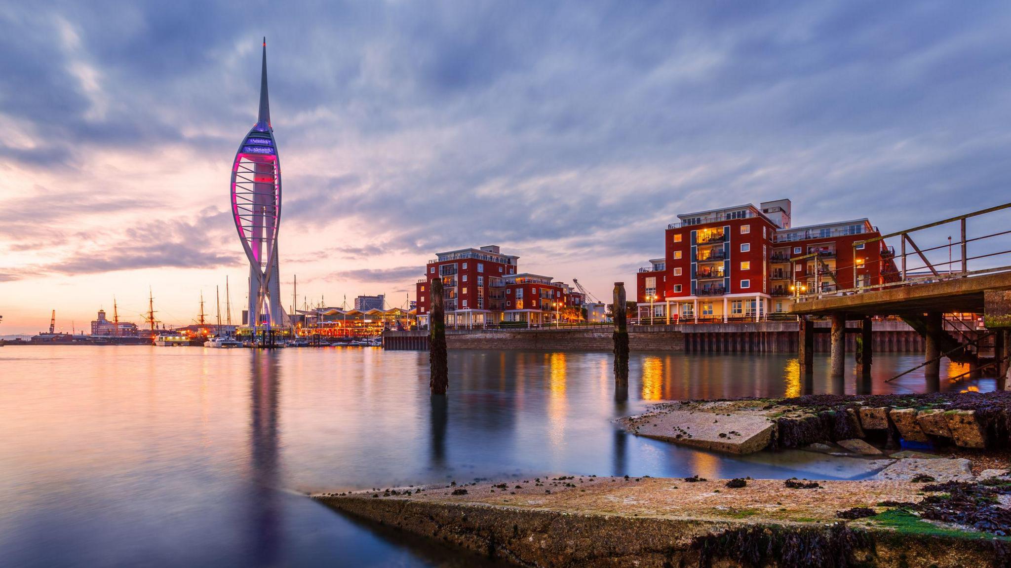 Dusk view of Portsmouth waterfront with the Spinnaker Tower lit in purple and white, surrounded by modern red-brick buildings and calm water reflecting city lights.