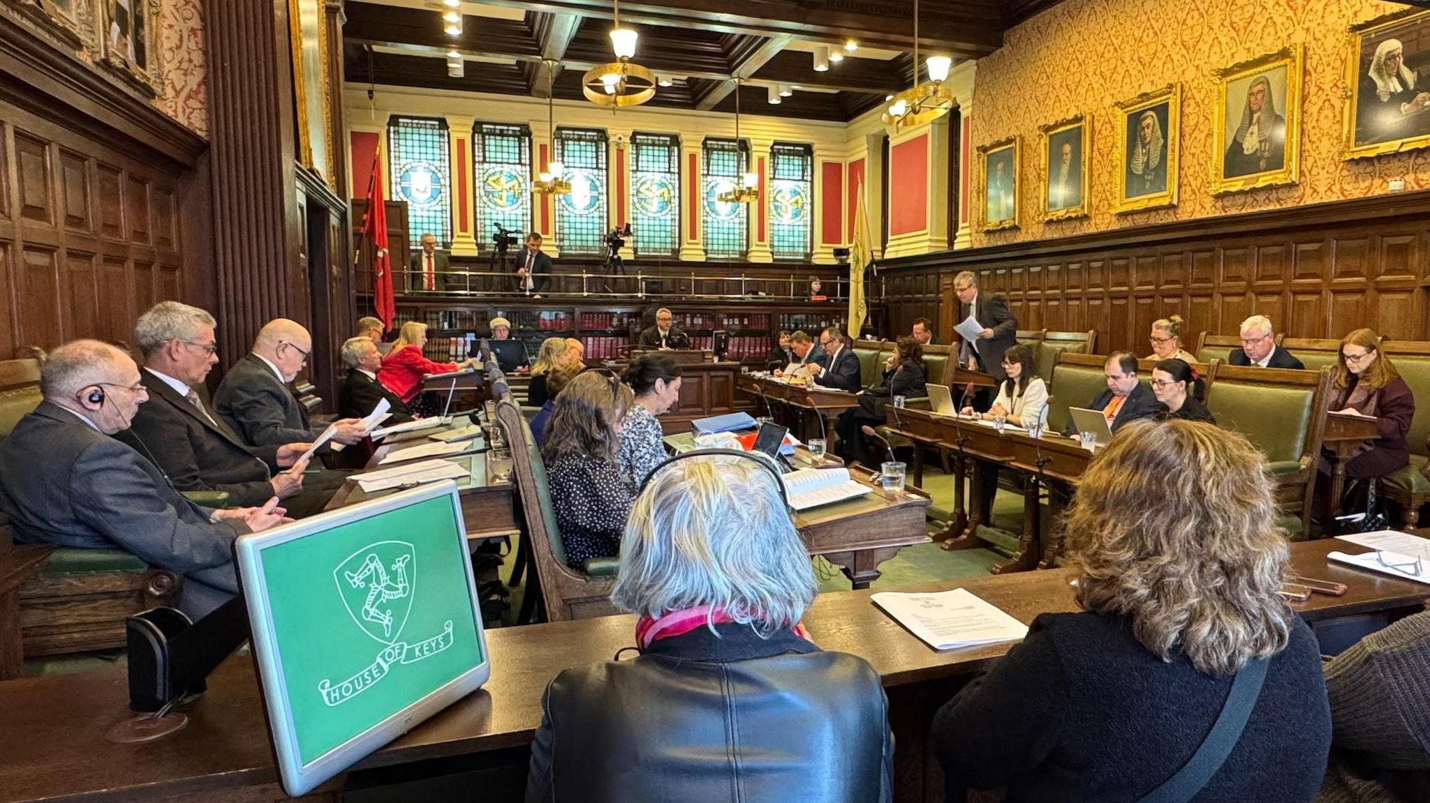 MHKs debating the bill in the House of Keys Chamber, with people sitting in the public gallery at the fore. The camber has wooden panels on the walls and stained glass windows at the back and the members are sitting on green leather clad seats at rows of wooden benches on either side of the chamber.