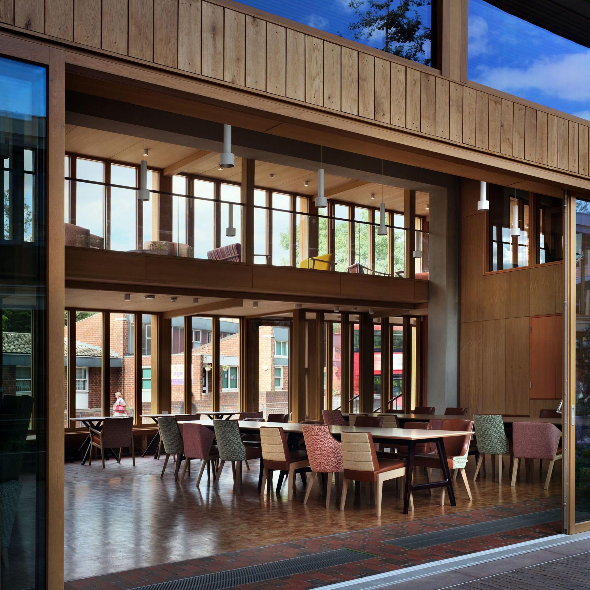 Looking in through large open glass doors to a double-height timber-walled communal room with a large table and chairs of different colours
