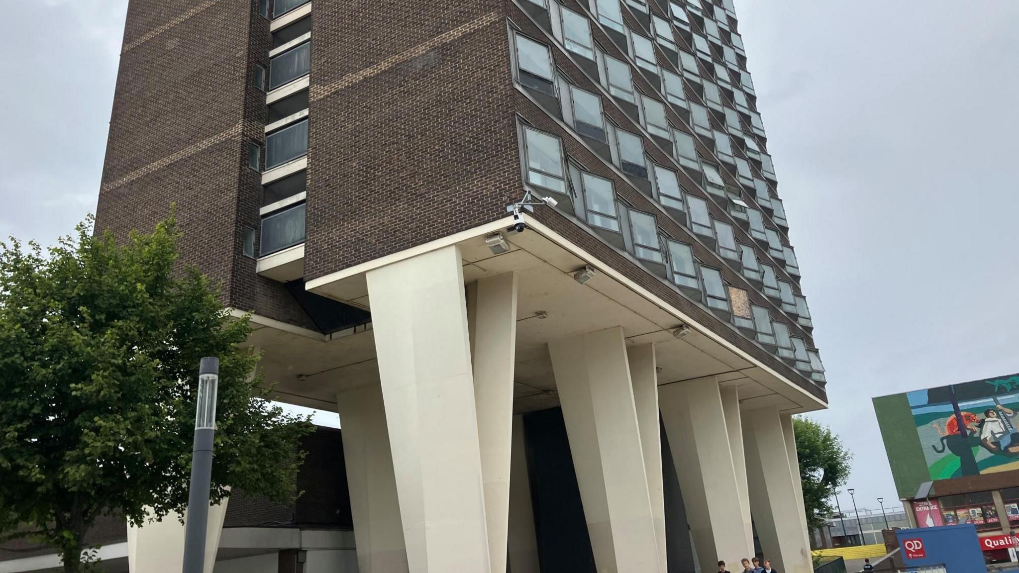 A tower block is pictured standing on large, cream-coloured supporting stilts, which are V-shaped. Rows of windows are seen on the different floors of the tower block.