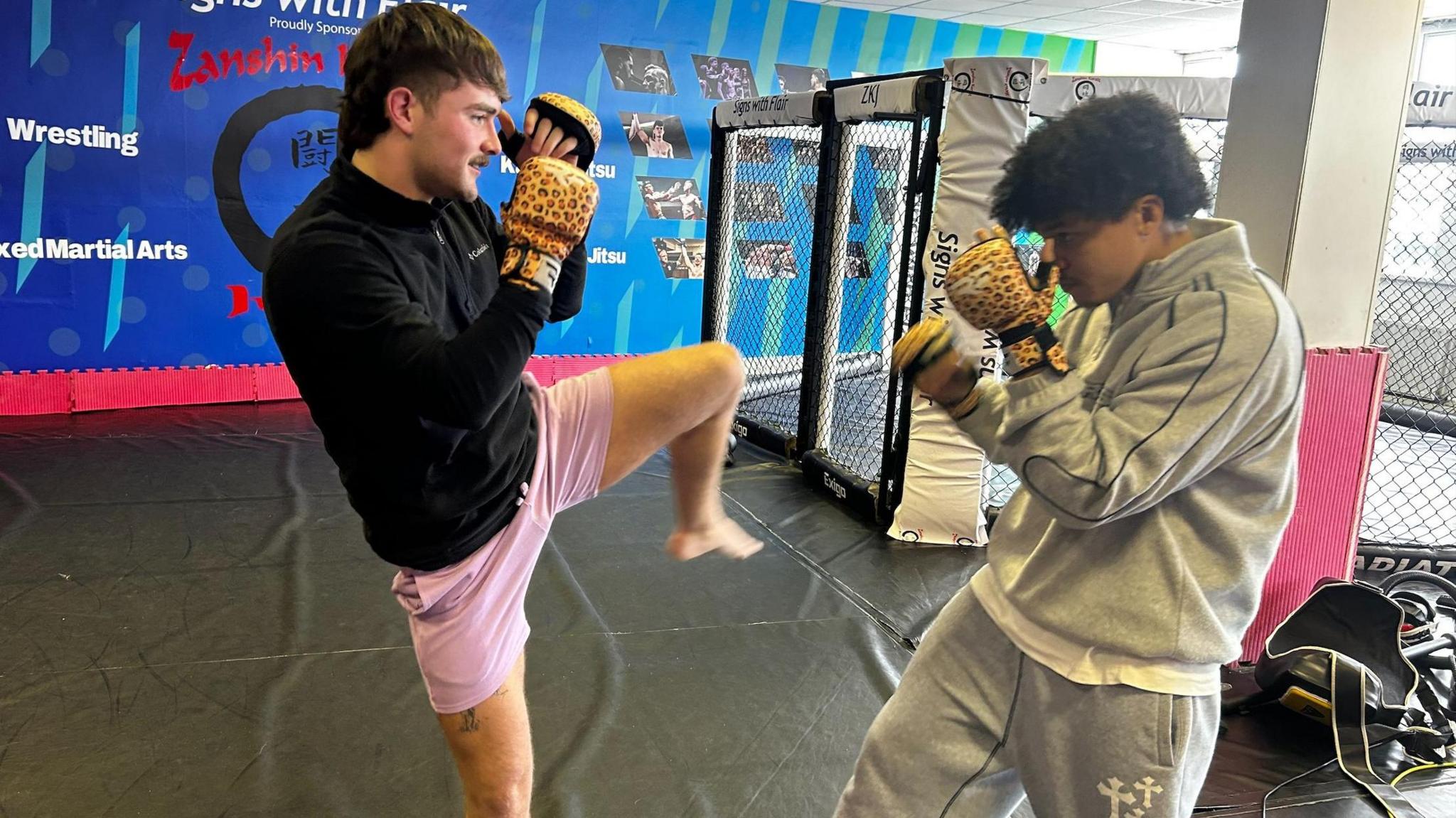 A young man in pink shorts and a black top spars with a young man in a grey tracksuit. They are both wearing leopard print gloves.