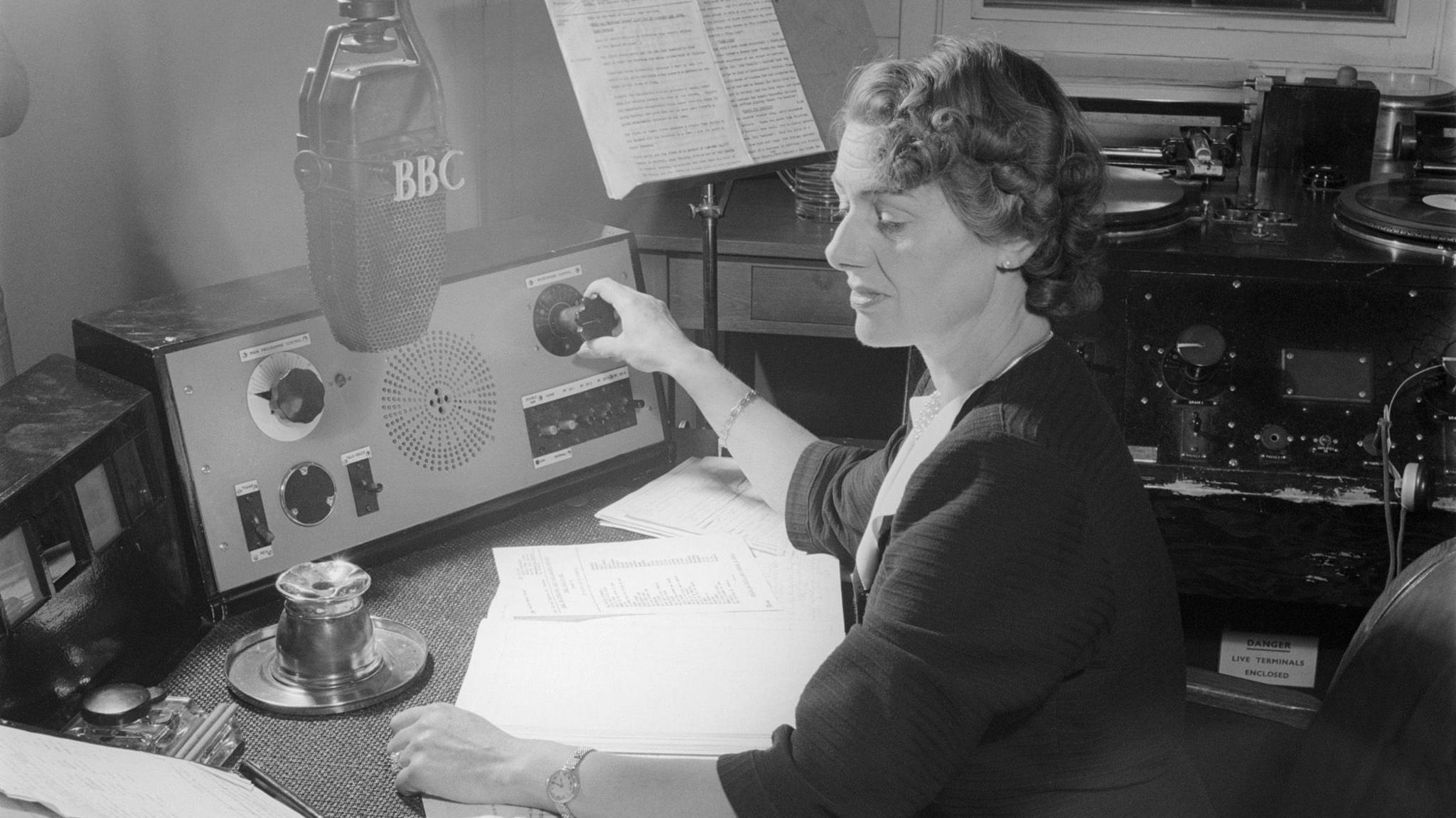 A black and white picture of a woman sitting at a broadcasting desk in front of a microphone which says BBC on it. She is surrounded by old recording equipment.