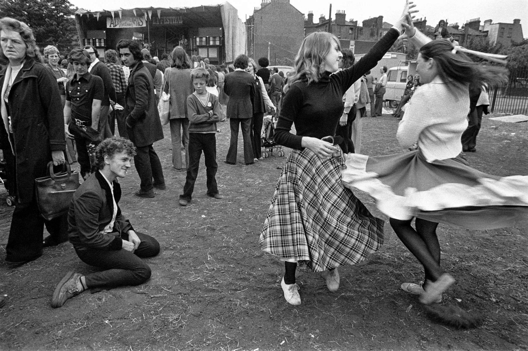 A black and white image of two girls dancing at an outdoor music event while a young man sitting on the ground looks on. 