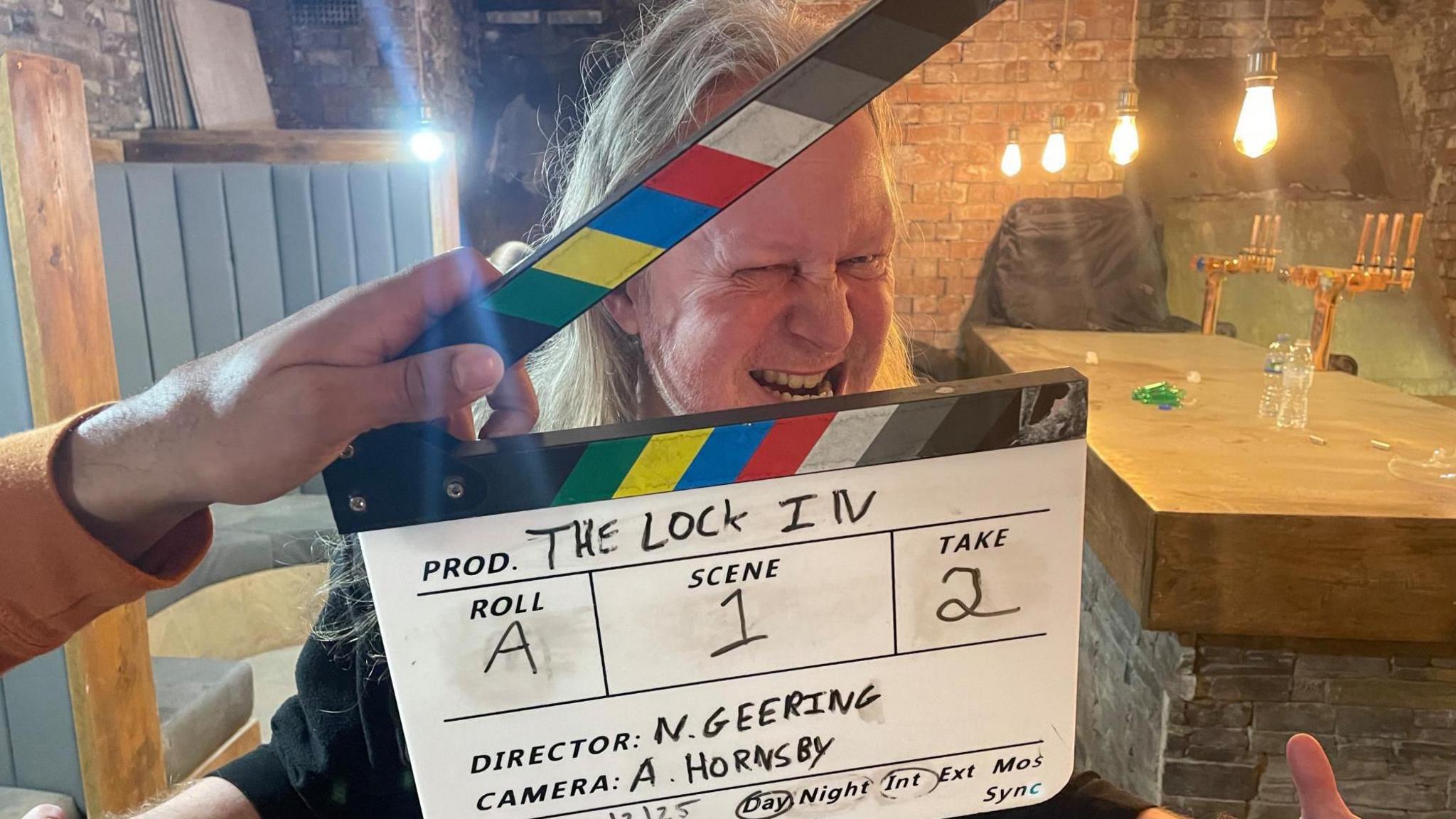 A clapperboard is held up infront of the face of a man with long white hair and gritted teeth