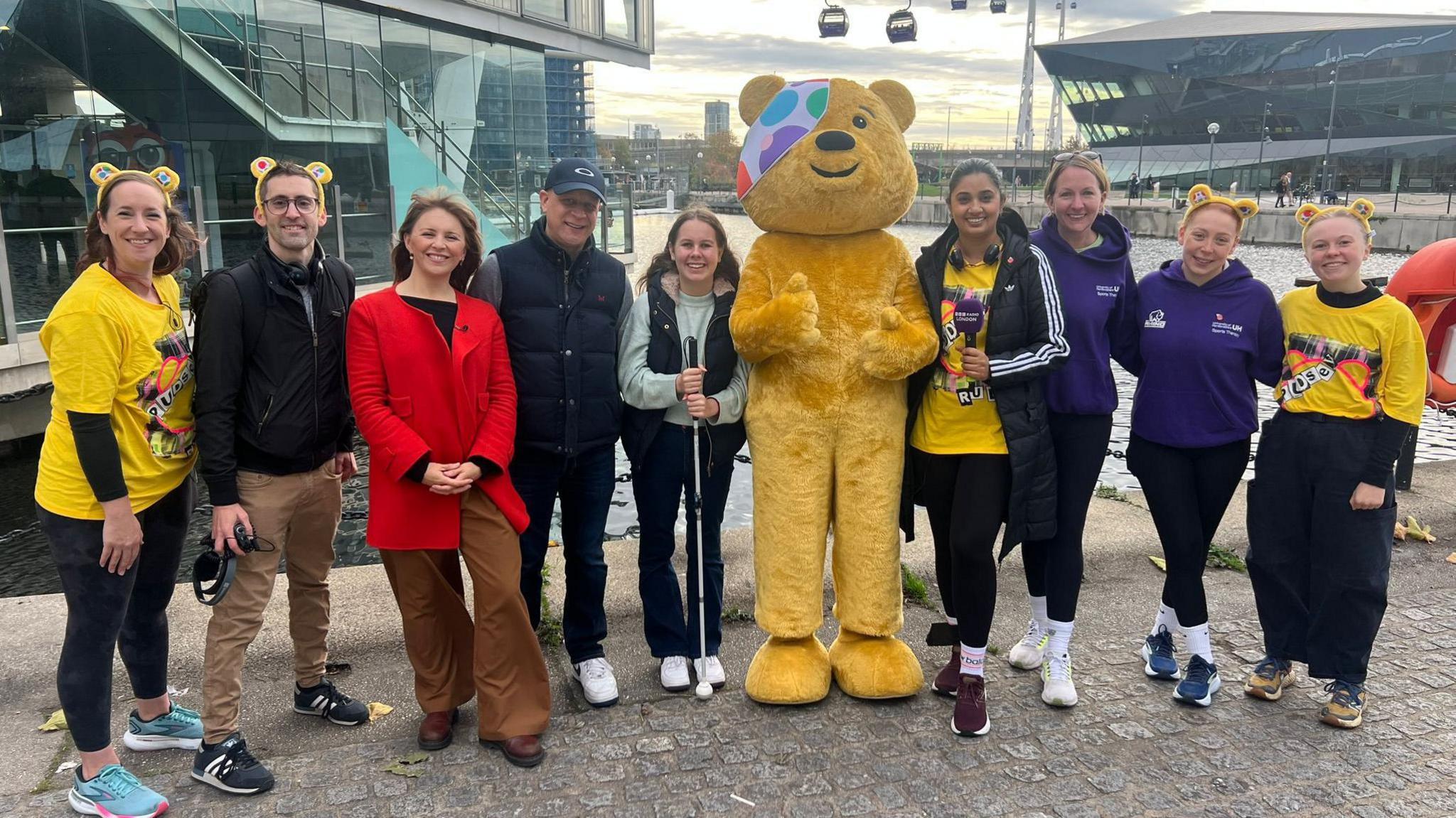 A big yellow bear puts his thumbs up while standing in the middle of a group of people by London's cable car, with the pods in the background. Eleanor is with her dad, Tim and is holding her white cane.